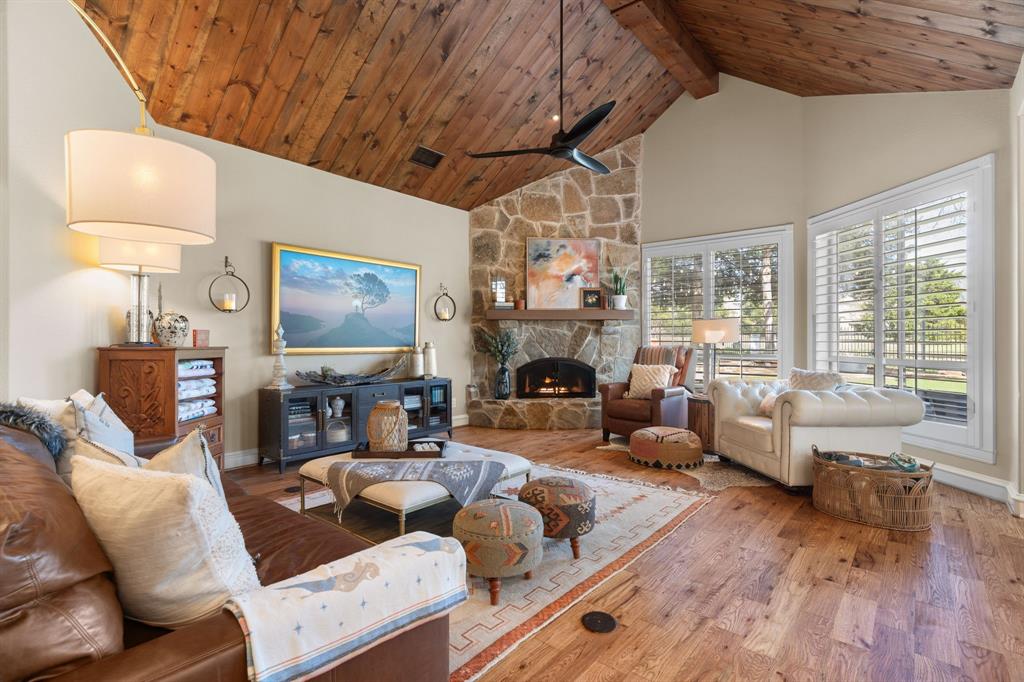 2404 Racheal Drive Heath, TX 75032 - Photo 10 of 40 The living room is a true showpiece, rich in character and warmth, featuring hardwood floors and a dramatic floor-to-ceiling stone gas fireplace with a raised hearth, wood mantel, and a new custom iron fireplace cover. Two expansive windows with plantation shutters frame serene backyard views, while a striking vaulted pine ceiling with a prominent center beam and a new modern aviator-style ceiling fan adds architectural distinction.