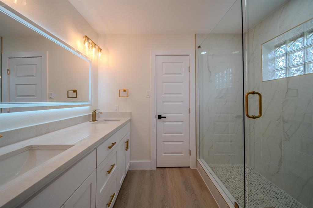 142 Ridgegate Drive Garland, TX 75040 - Photo 22 of 26 Contemporary bathroom featuring a spacious glass-enclosed shower with a glass block window, a dual vanity with undermount sinks and light countertops, a large back-lit mirror, and wood-finish flooring