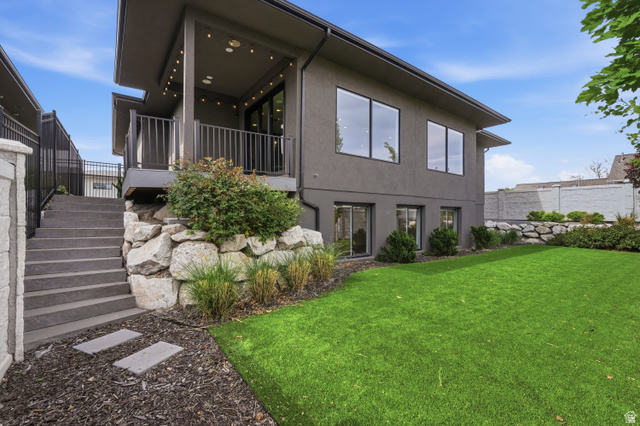 653 East Vandalay Lane Draper, UT 84020 - Photo 45 of 61 Contemporary grey exterior featuring a spacious elevated deck with string lighting, a manicured lawn, an integrated rock retaining wall, and a concrete staircase with stone accents