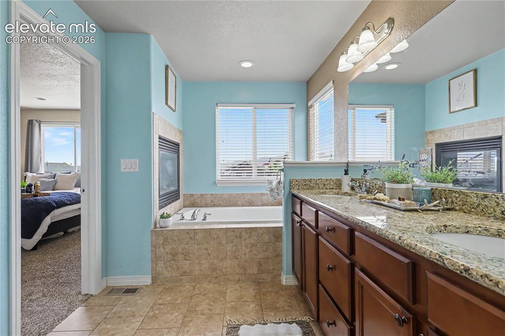 6598 Alliance Loop Colorado Springs, CO 80925 - Photo 19 of 39 Bathroom featuring a dual vanity with granite countertops and undermount sinks, a built-in fireplace, a jetted tub, and tiled flooring
