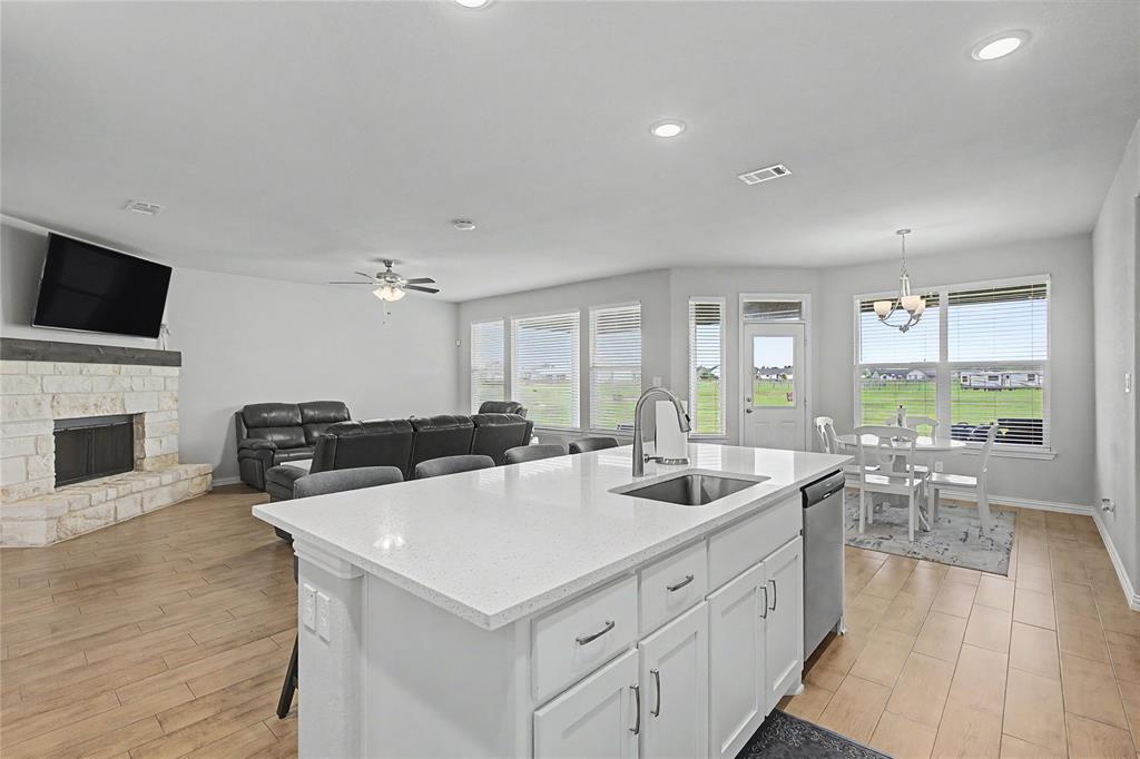 Expansive kitchen island with a white countertop and undermount sink, stainless steel dishwasher, wood-finish flooring, stone fireplace, and ample natural light from multiple windows