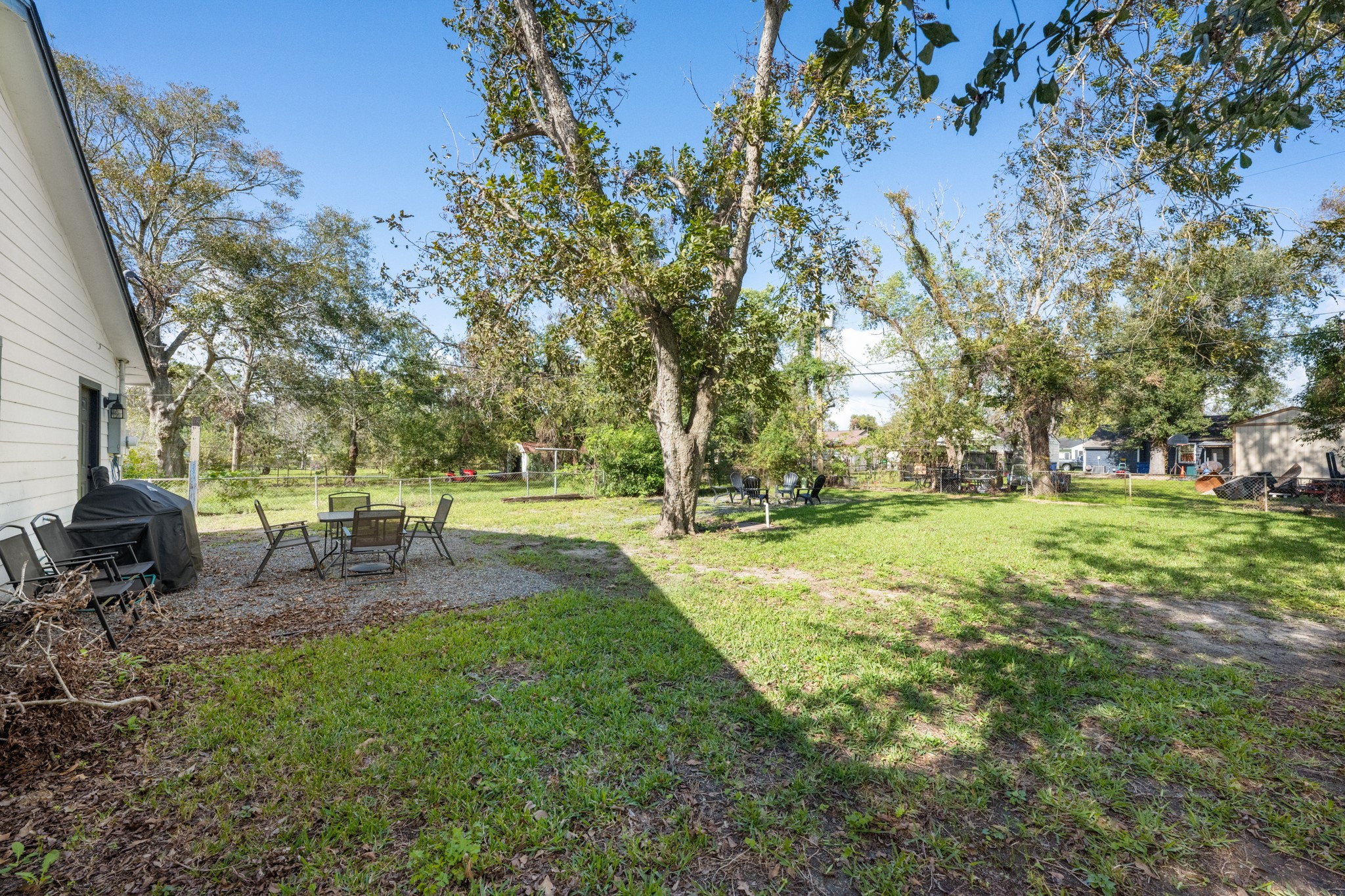 729 Kyle Angleton, TX 77515 - Photo 34 of 36 The photo showcases a spacious backyard with mature trees providing shade. There's a patio area with outdoor seating and a grill, offering a comfortable space for relaxation or entertaining. The yard is grassy and well-maintained, ideal for family activities or gardening.