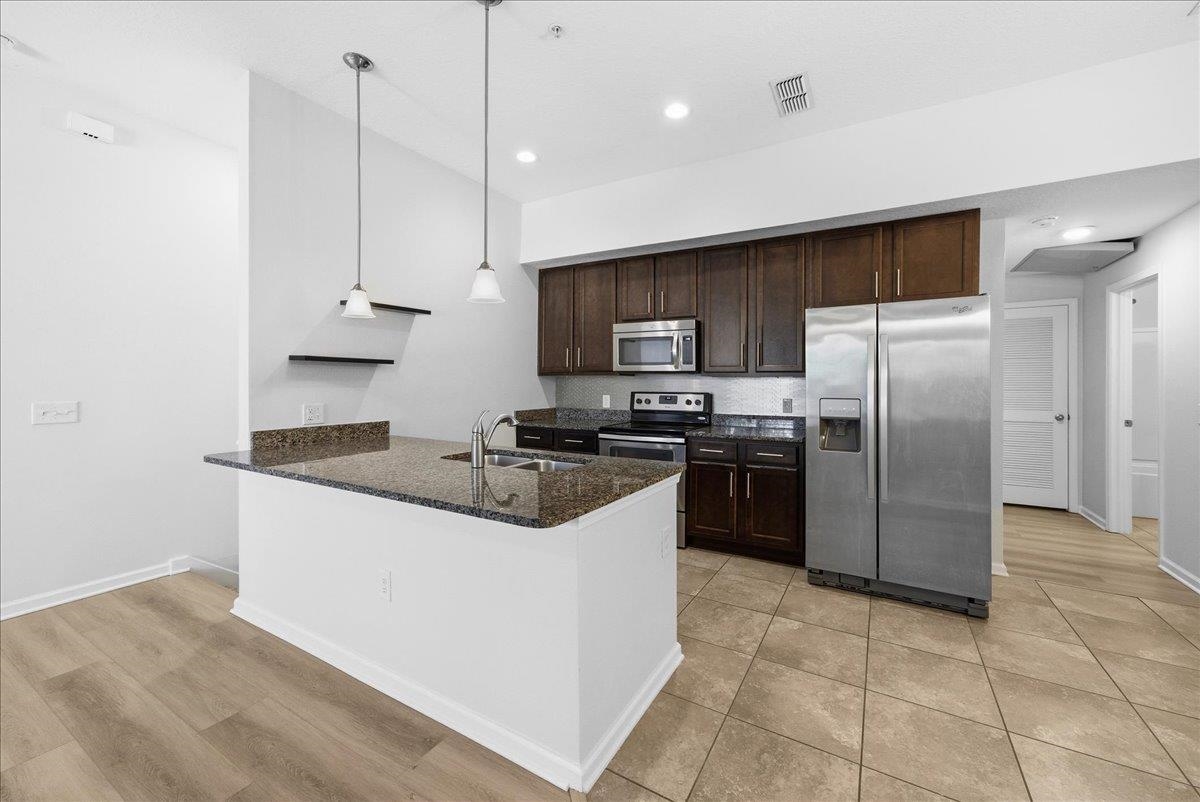 Kitchen with appliances with stainless steel finishes, dark brown cabinetry, dark stone countertops, a peninsula, and decorative light fixtures