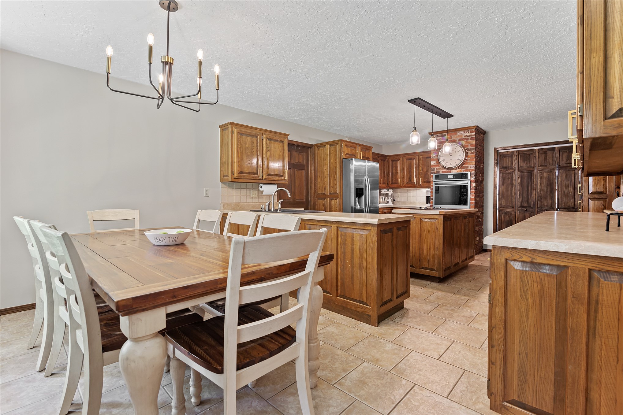 9644 19th Road Nowata, OK 74048 - Photo 5 of 36 Open-concept kitchen and dining area featuring tile flooring, wood cabinetry, built-in oven, stainless steel refrigerator, and a brick accent wall with a large clock