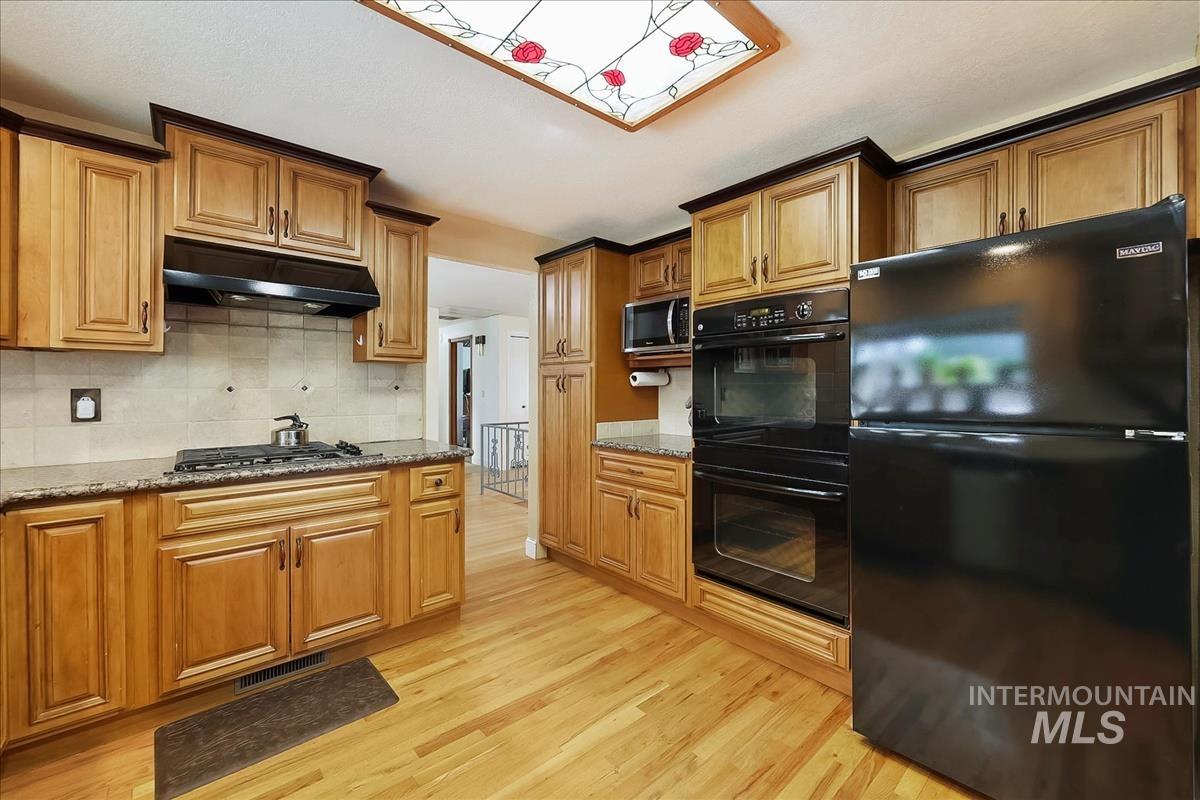 11875 West Hidden Valley Rim Road Boise, ID 83709 - Photo 9 of 36 Kitchen with black appliances, dark stone countertops, wood finish cabinetry, light wood-style flooring, and decorative backsplash