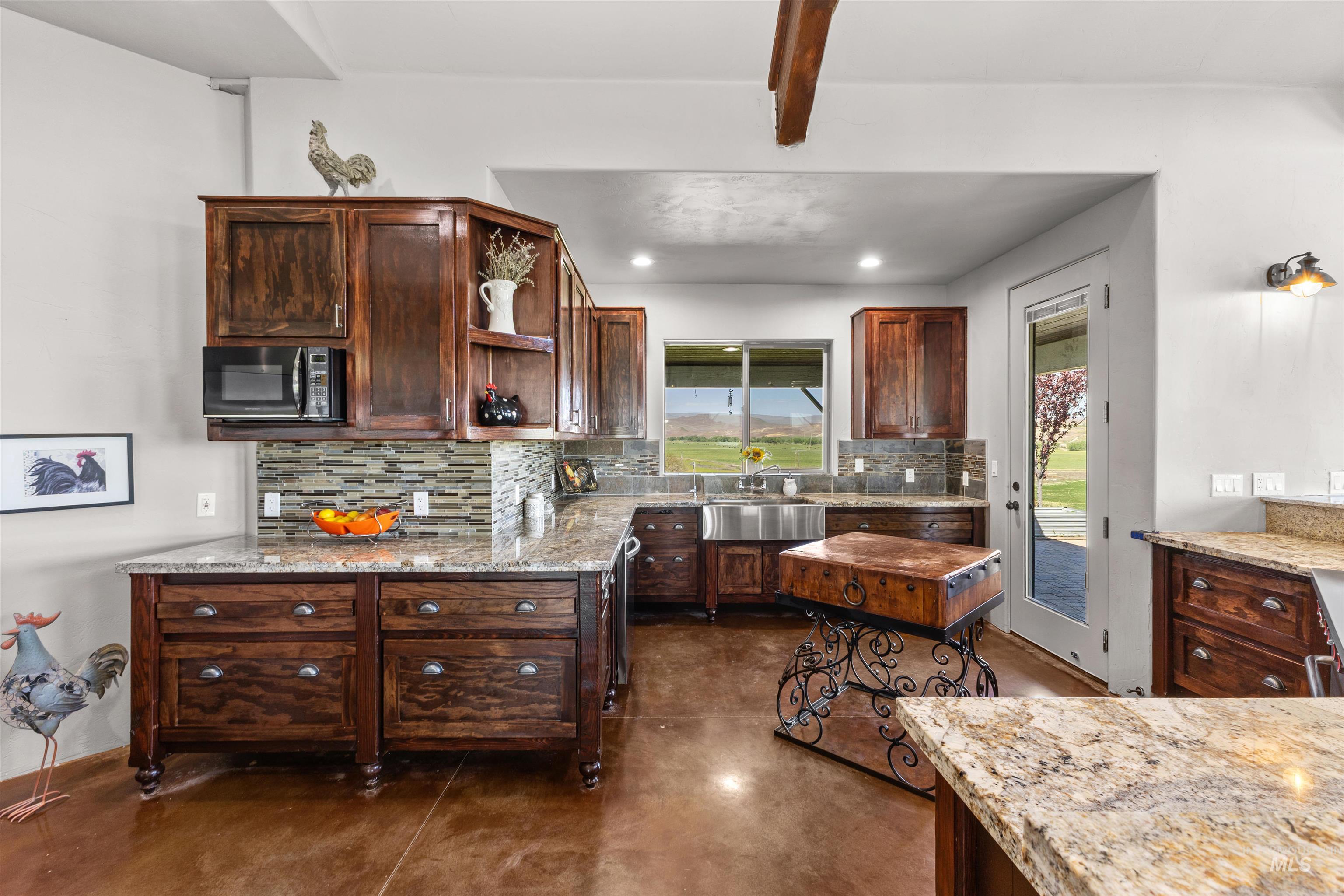 1030 Desert Glen Road Adrian, OR 97901 - Photo 26 of 47 Kitchen featuring extensive dark wood cabinetry, light-toned granite countertops, a stacked stone tile backsplash, a stainless steel farmhouse sink, and a glass-paneled door to the exterior