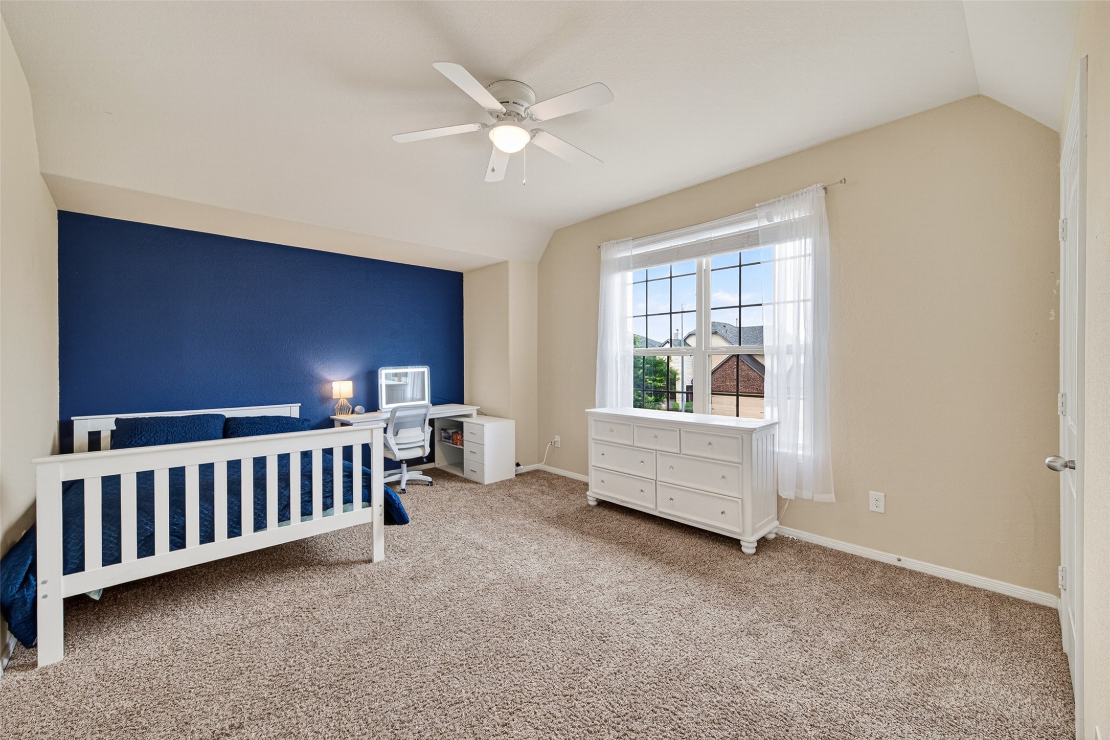 25611 Ellis Ridge Court Katy, TX 77494 - Photo 32 of 49 This bright bedroom features a cozy carpet, a bold blue accent wall, and a large window with sheer curtains, offering ample natural light. There’s a ceiling fan, a white bed, a desk, and a dresser, creating a functional and inviting space.