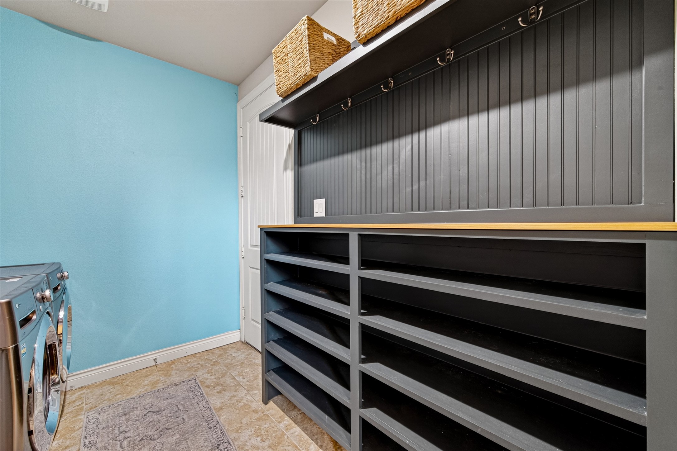 25611 Ellis Ridge Court Katy, TX 77494 - Photo 19 of 49 This photo showcases a laundry room with a bright blue accent wall and tiled flooring. It features a modern washer and dryer, ample open shelving for storage, and wall hooks for hanging. A practical and organized space for household chores.