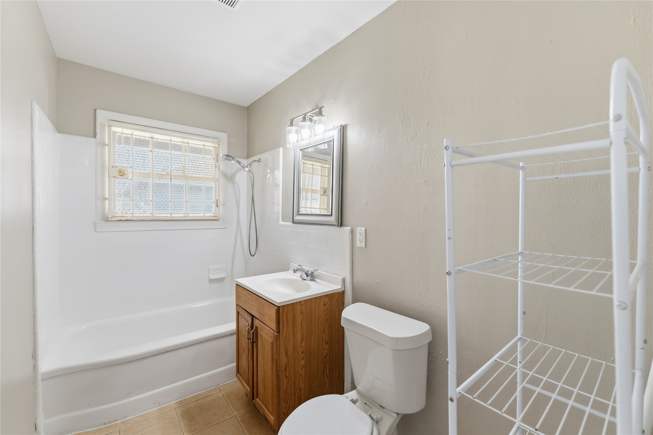 4814 Burma Road Houston, TX 77033 - Photo 14 of 16 Bathroom with a neutral palette, featuring a bathtub, shower, and a window for natural light. It includes a wooden vanity with a mirror, a toilet, and a white shelving unit for storage.