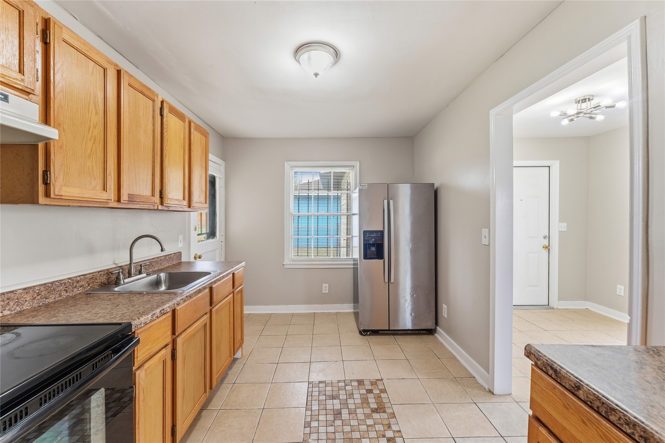 4814 Burma Road Houston, TX 77033 - Photo 5 of 16 Kitchen features light wood cabinets, granite-style countertops, and a neutral tile floor. It includes a stainless steel refrigerator and a window for natural light. An adjacent room is visible through a doorway, enhancing the open feel.