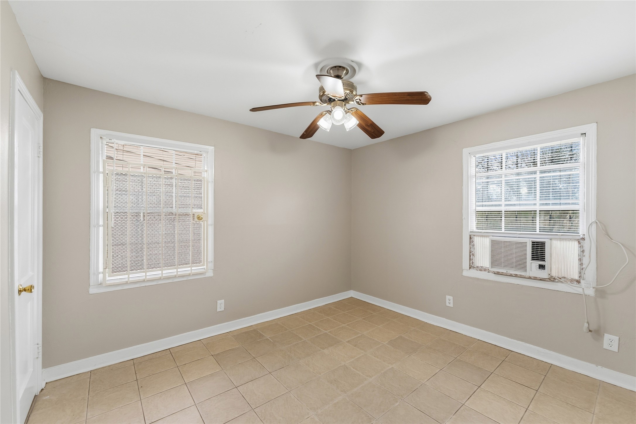 4814 Burma Road Houston, TX 77033 - Photo 10 of 16 This room features neutral-colored walls, tiled flooring, and two windows with blinds. It includes a ceiling fan with lights and a window-mounted air conditioning unit. The space is well-lit and suitable for various uses.