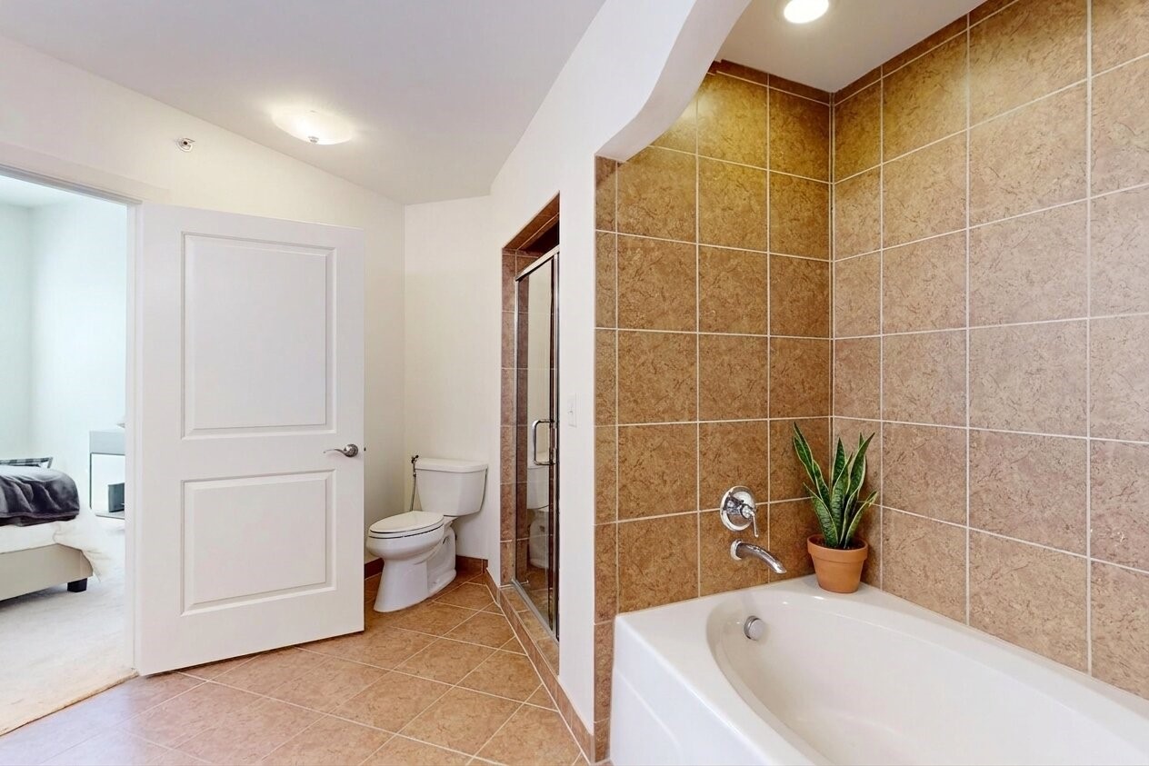 1010 Rosine Street, Unit 29 Houston, TX 77019 - Photo 8 of 16 This modern bathroom features a spacious tub with stylish brown tile surround, a separate glass-enclosed shower, and a sleek white toilet. A potted plant adds a touch of greenery, and an open door leads to a bright adjoining bedroom. (Virtual Staging)