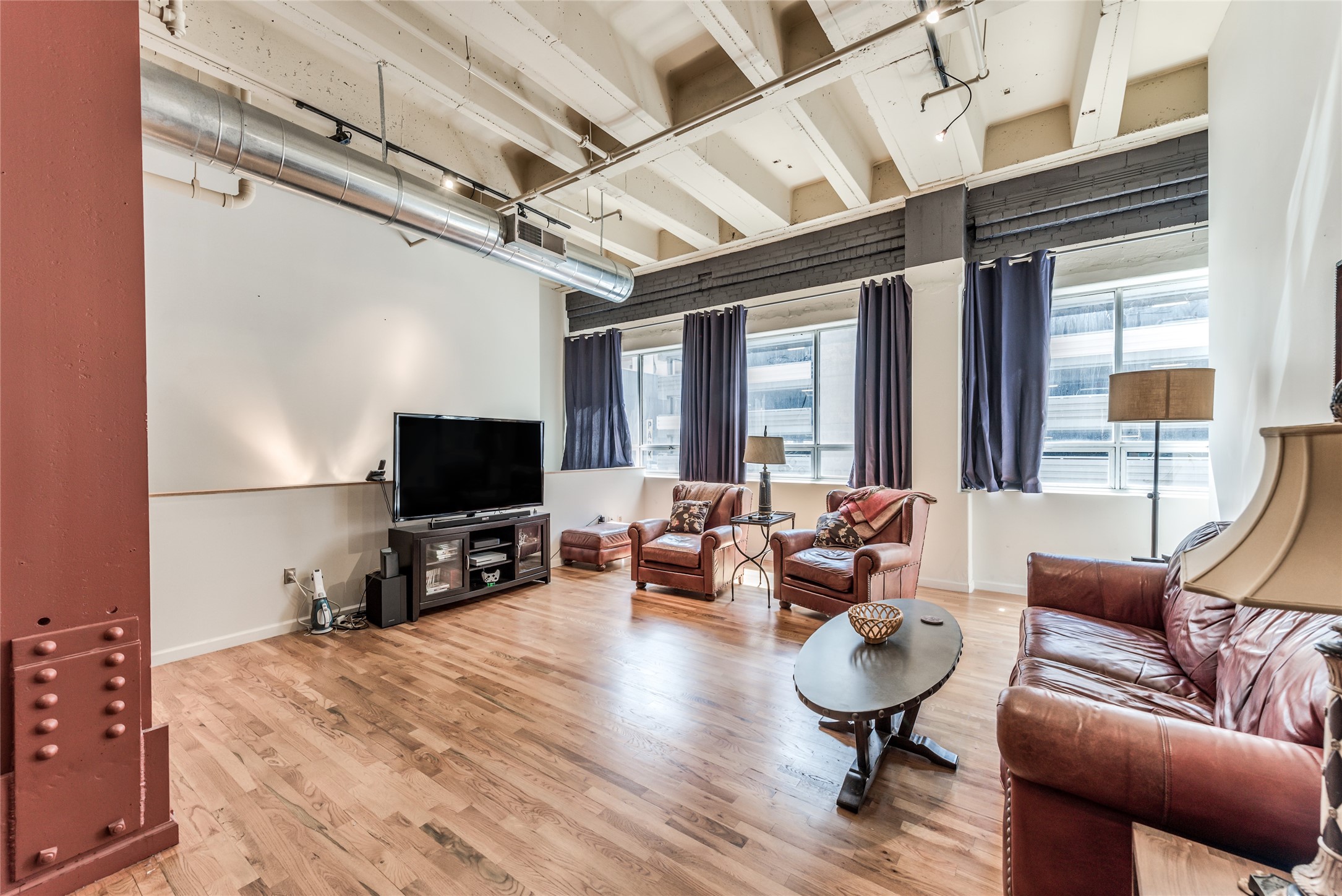 705 Main Street, Unit 215 Houston, TX 77002 - Photo 7 of 36 This photo showcases a spacious, industrial-style living room with high ceilings, exposed beams, and ductwork. It features large windows, hardwood floors, leather furniture, and a cozy seating area with a TV. The space is bright and modern, ideal for comfortable living.