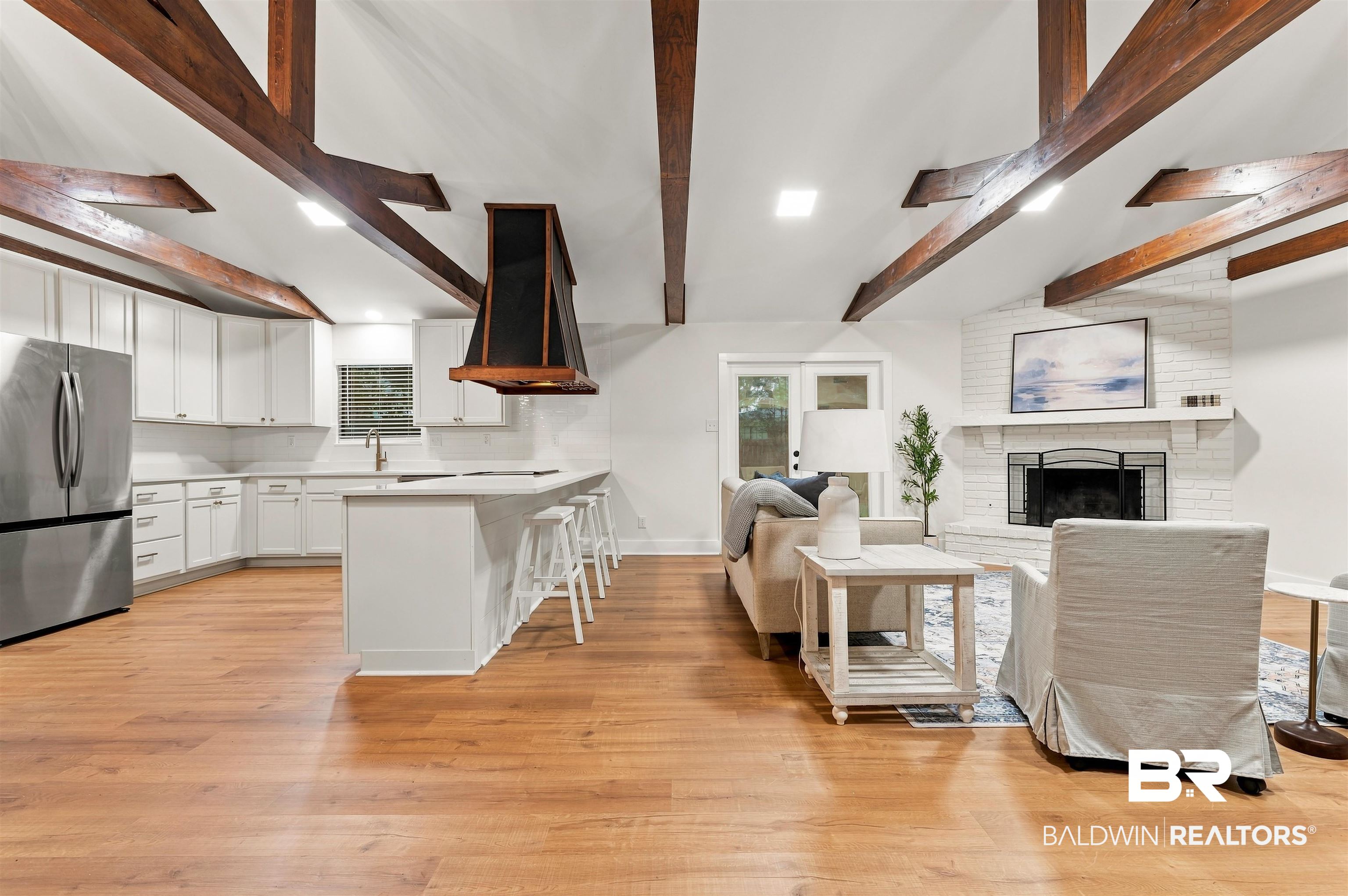 599 Ridgewood Drive Daphne, AL 36526 - Photo 6 of 41 Open-concept living space featuring vaulted ceilings with exposed wood beams, wood-finish flooring, a white brick fireplace, a kitchen island with seating, and stainless steel appliances