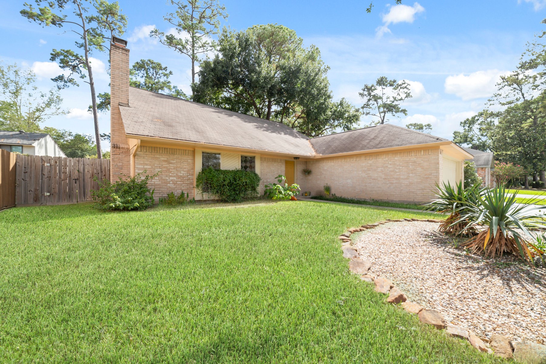 25402 Mill Pond Lane Spring, TX 77373 - Photo 6 of 24 This photo showcases a charming single-story brick home with a large, well-maintained front lawn. The property features a wooden fence, mature trees, and a landscaped area with rocks and plants, offering a peaceful suburban setting.