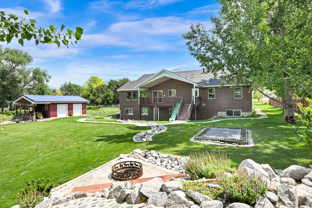13658 South Fort Street Draper, UT 84020 - Photo 65 of 87 Expansive green lawn featuring a built-in fire pit area with stone landscaping, a detached two-car garage with an attached covered porch, a two-story brick exterior, and a second-story balcony