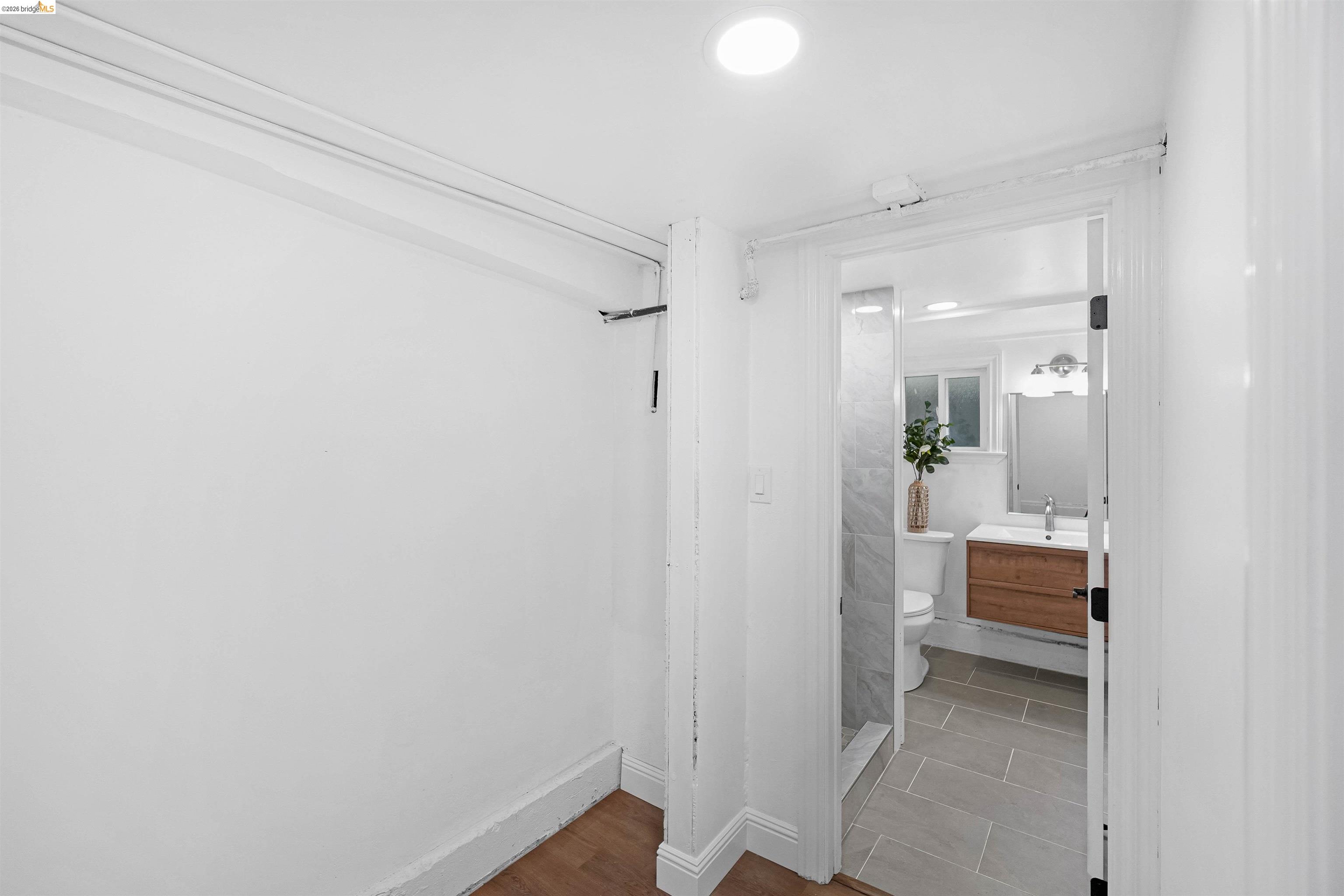 1812 Woolsey Street Berkeley, CA 94703 - Photo 40 of 48 White-painted interior with wood-finish flooring, opening to a bathroom featuring a wood vanity with a white countertop, a shower with stone-look tile, and grey tile flooring