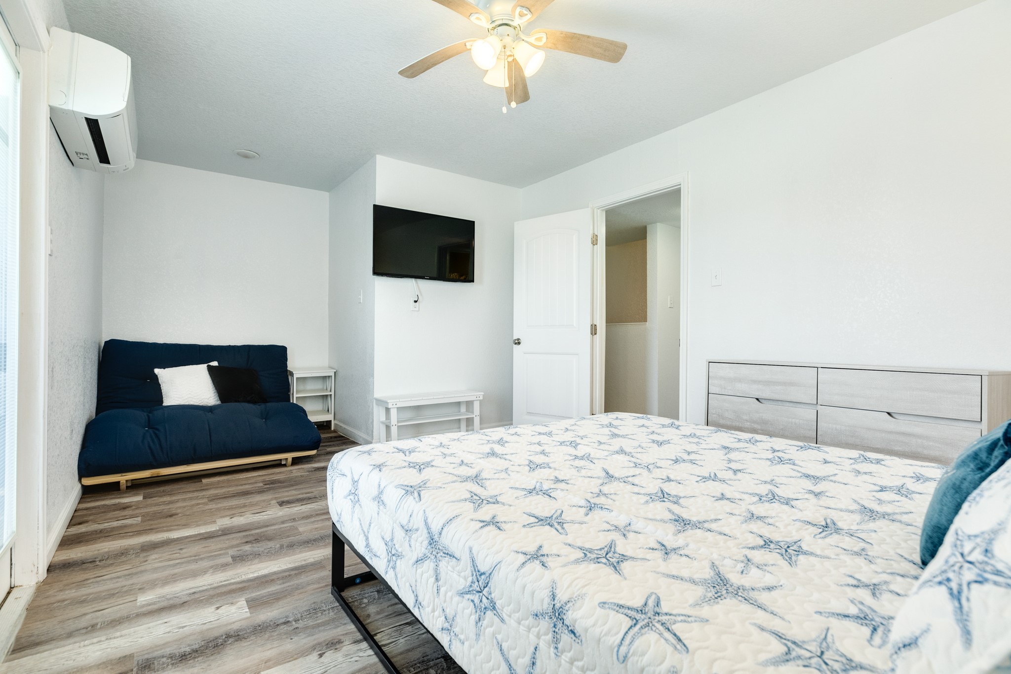 101 Pelican Place, Unit 7 Freeport, TX 77541 - Photo 21 of 44 This bright bedroom features a ceiling fan, air conditioning, and wood-style flooring. It offers a comfortable layout with a bed, a blue futon, a flat-screen TV, and a dresser, creating a cozy and functional space.