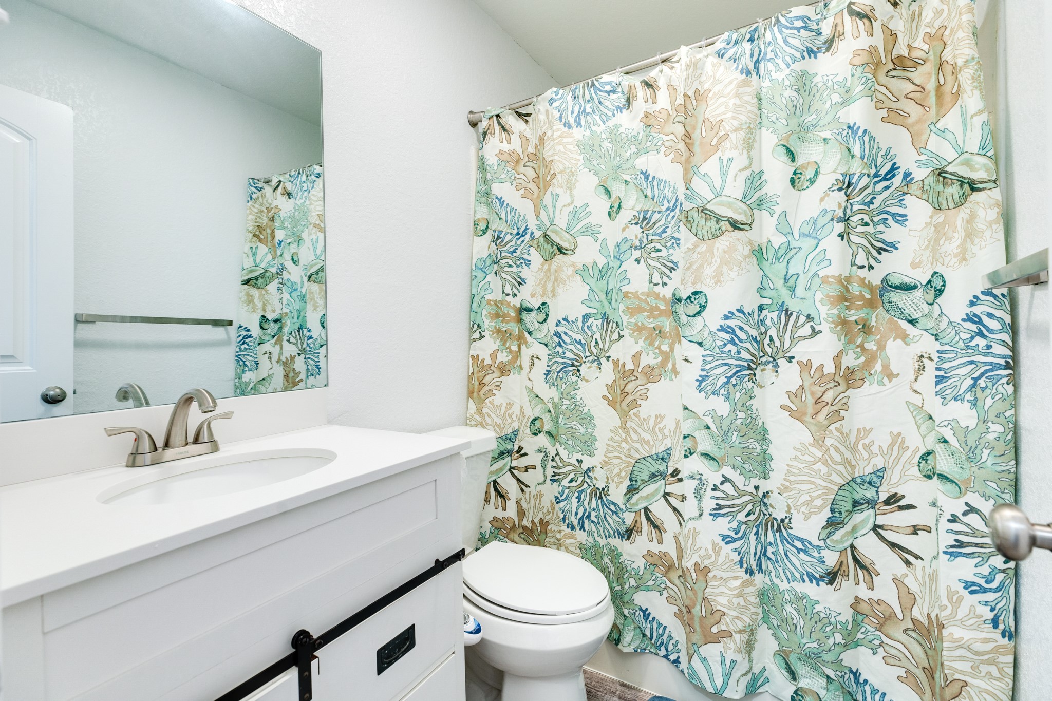101 Pelican Place, Unit 7 Freeport, TX 77541 - Photo 25 of 44 This is a bright bathroom featuring a white vanity with a modern faucet, a large mirror, and a toilet. The shower curtain has a colorful sea life design, adding a touch of coastal charm.