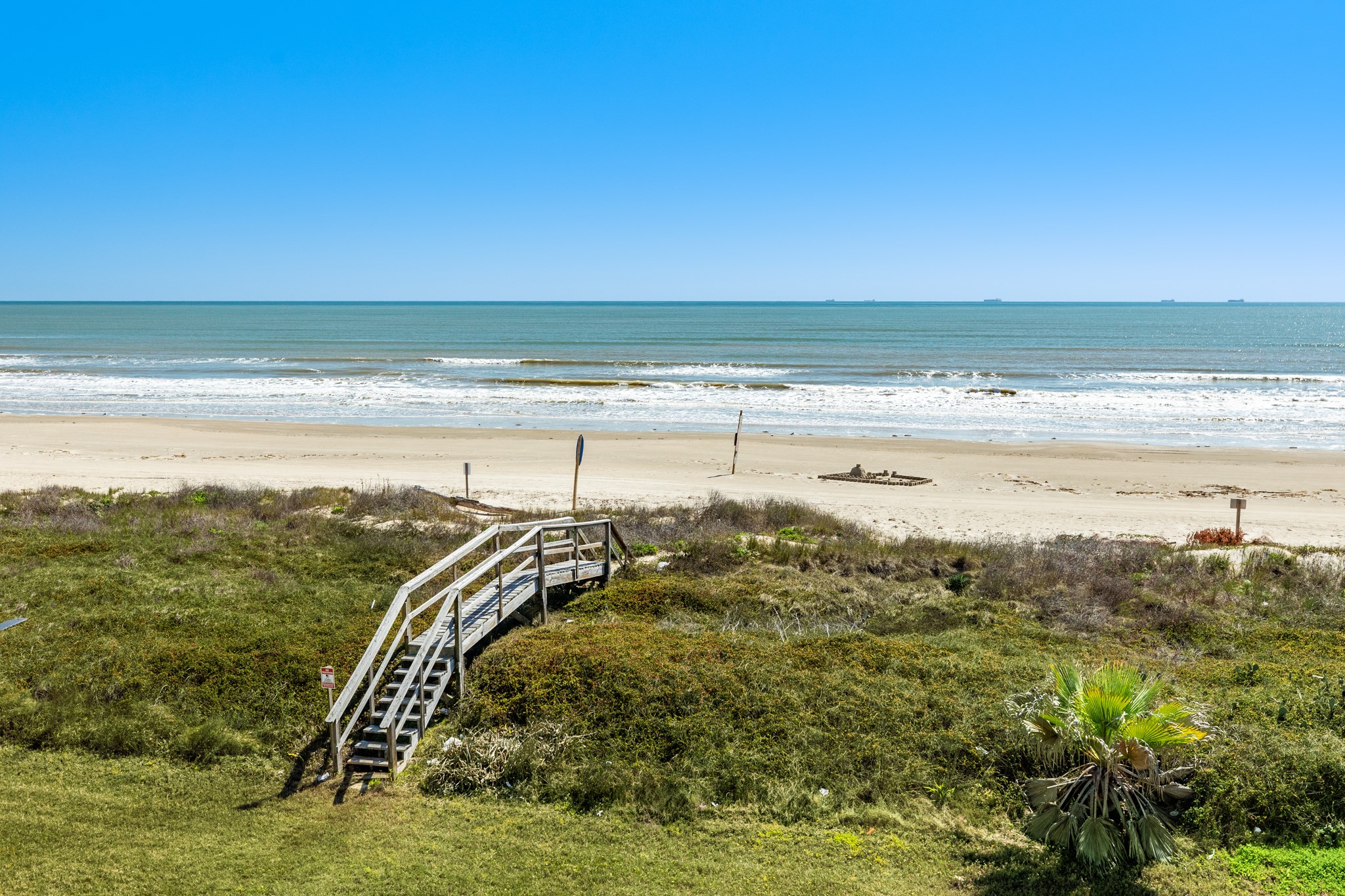 101 Pelican Place, Unit 7 Freeport, TX 77541 - Photo 34 of 44 This photo showcases a serene beachfront view with a wooden walkway leading to the sandy shore, ideal for those seeking a coastal home. The clear blue sky and gentle waves add to the tranquil setting.