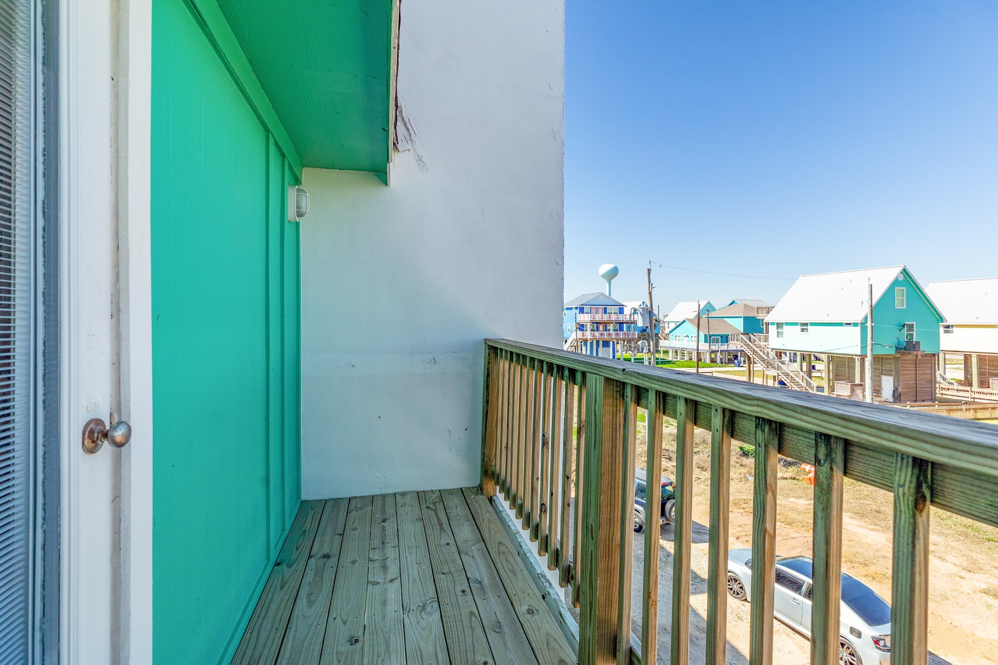 101 Pelican Place, Unit 7 Freeport, TX 77541 - Photo 35 of 44 This photo showcases a small wooden balcony with a green accent wall, offering views of a neighborhood with colorful houses and a clear blue sky. Ideal for enjoying fresh air and neighborhood sights.