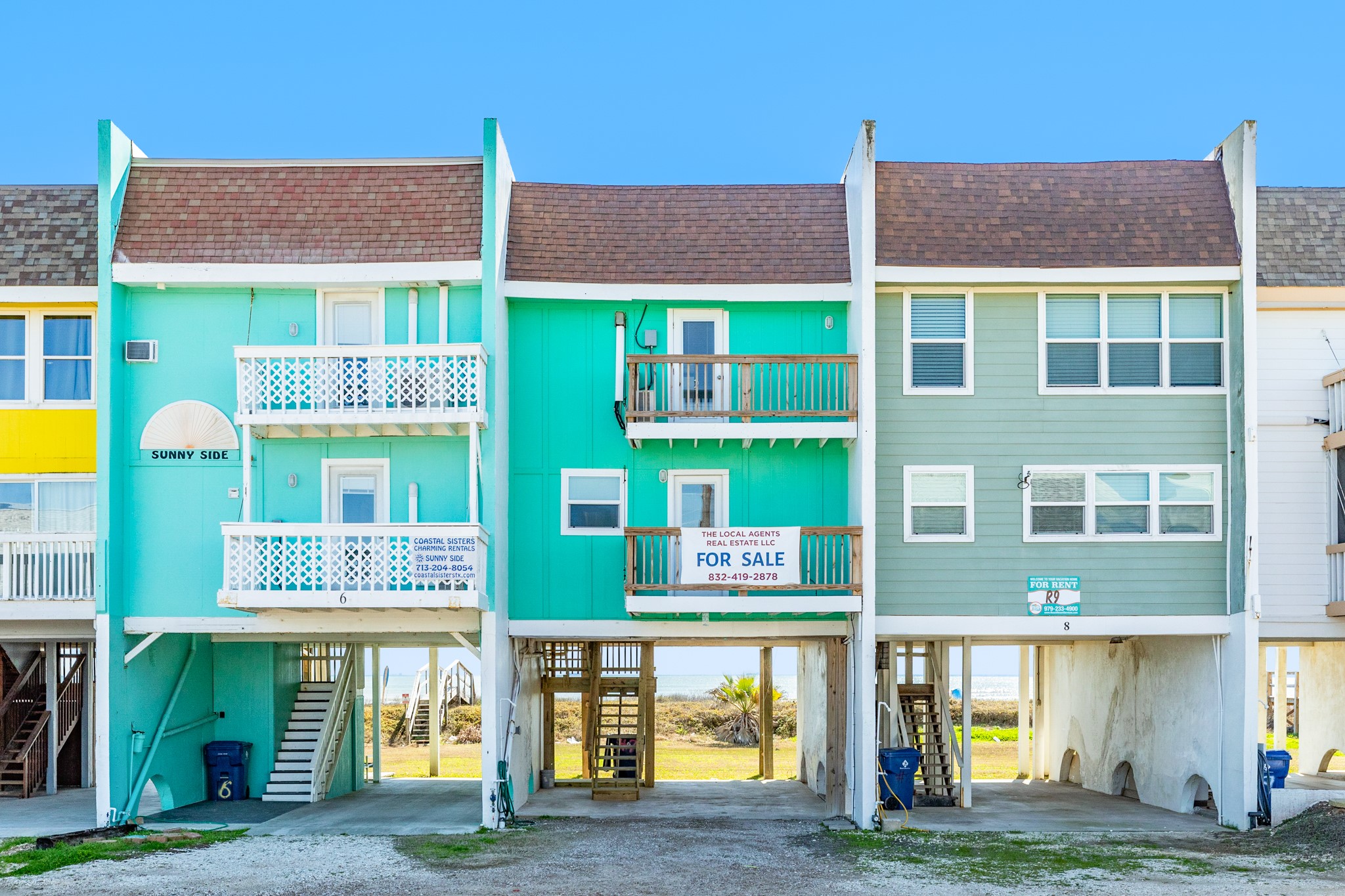 101 Pelican Place, Unit 7 Freeport, TX 77541 - Photo 5 of 44 This photo shows colorful, elevated beach houses with a bright teal unit prominently displayed. It features four decks and ample parking underneath. The property is coastal with a water view in the background, offering a seaside living experience.