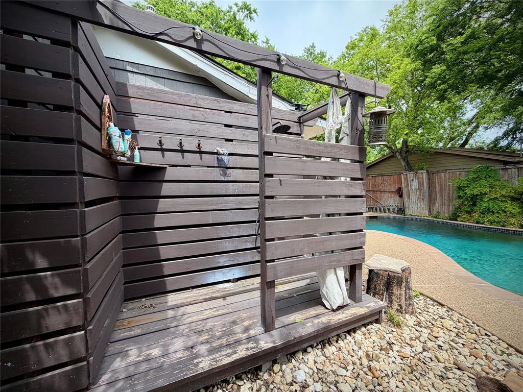 301 Forrest Avenue Cleburne, TX 76033 - Photo 3 of 14 Outdoor shower enclosure with slatted wood construction, integrated shelving, and wall-mounted hooks, positioned beside a freeform swimming pool with a concrete surround and pebble landscaping