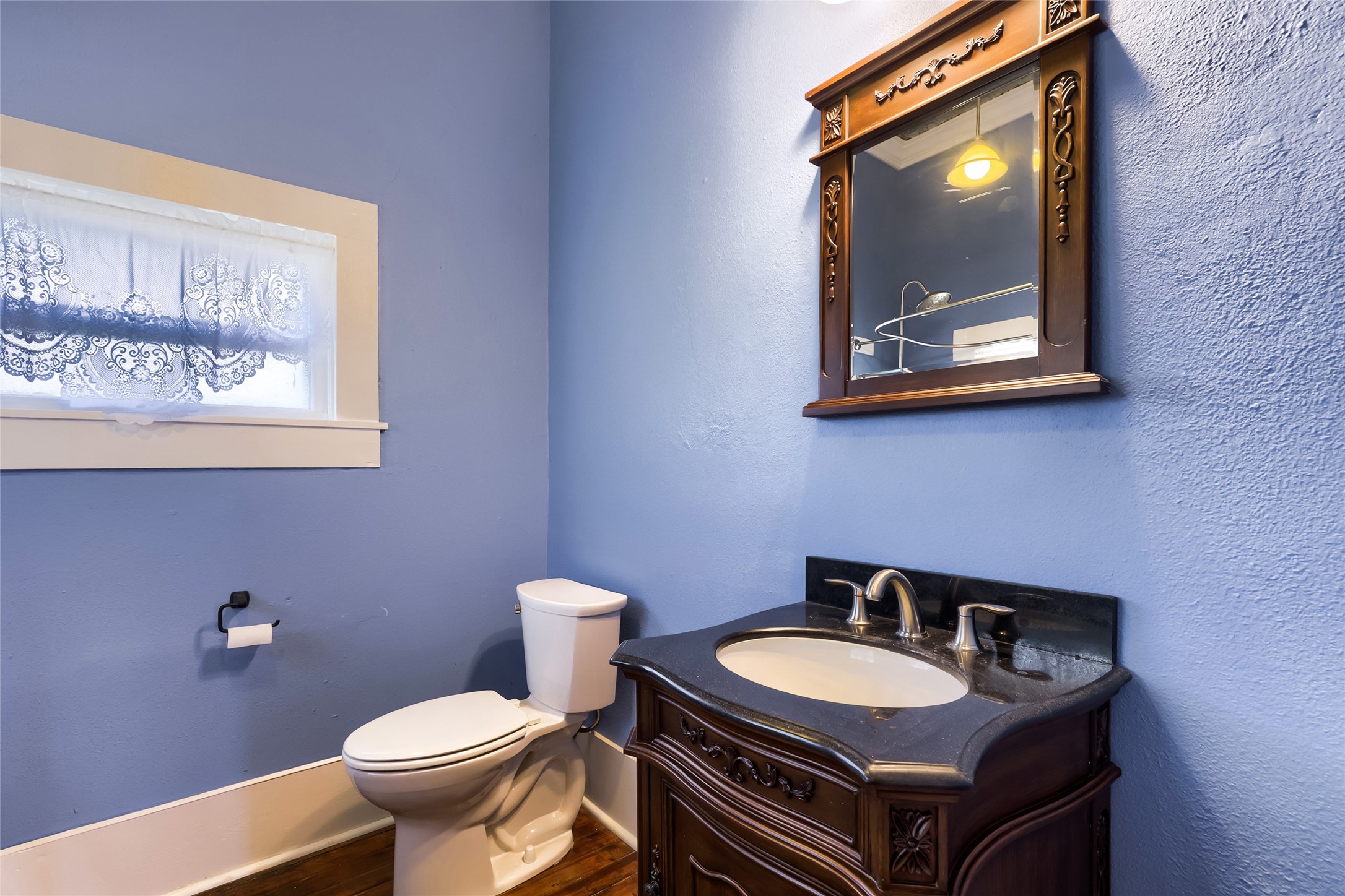 208 North Ave B Elgin, TX 78621 - Photo 15 of 29 Cozy bathroom with blue walls, featuring a vintage-style wooden vanity and mirror, a modern faucet, and a small window with decorative lace curtains.