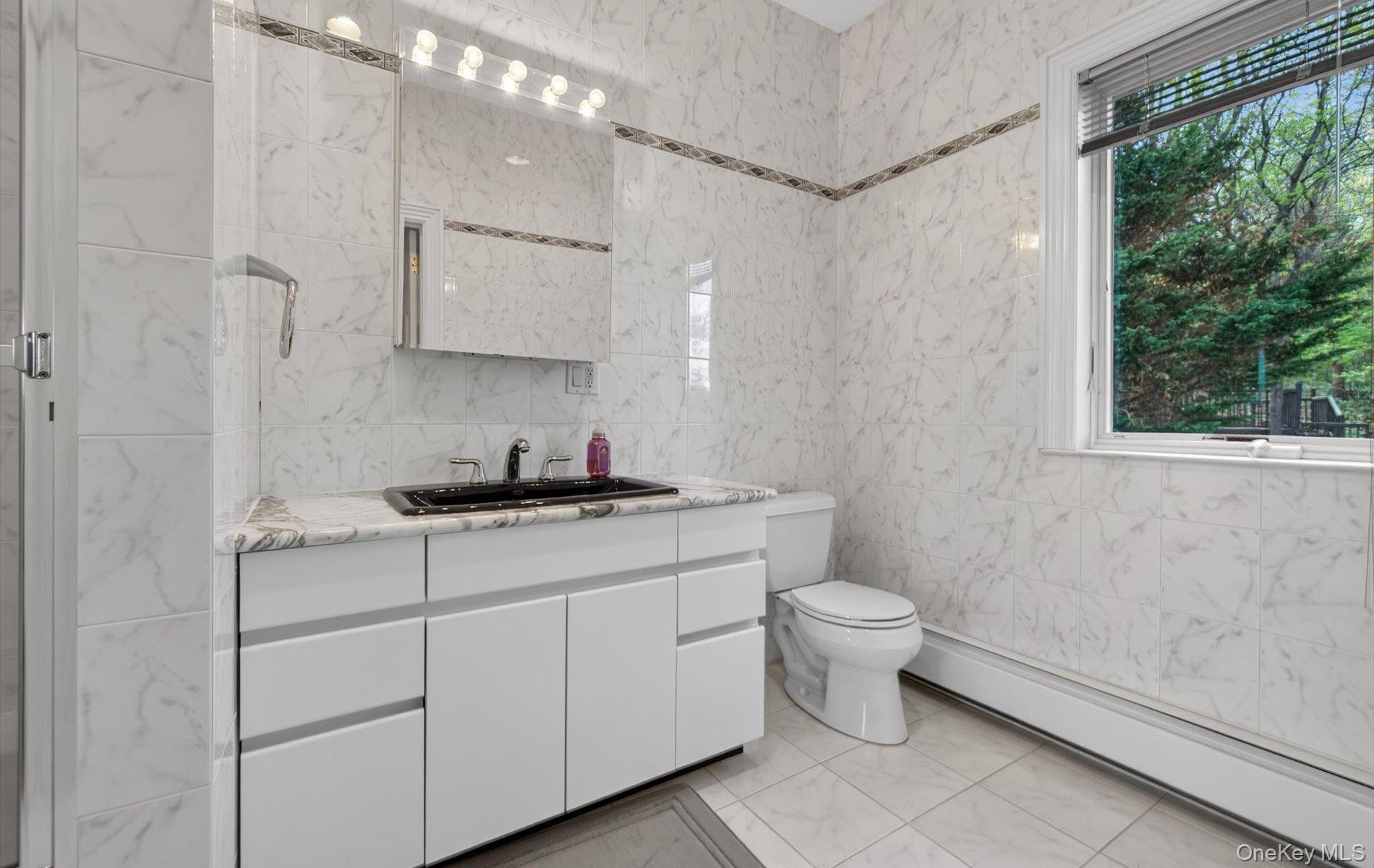 16 East Slope Road Bayville, NY 11709 - Photo 32 of 48 Bathroom featuring a white vanity with a black rectangular sink and chrome faucet, marble-look wall and floor tiling, and a window with blinds
