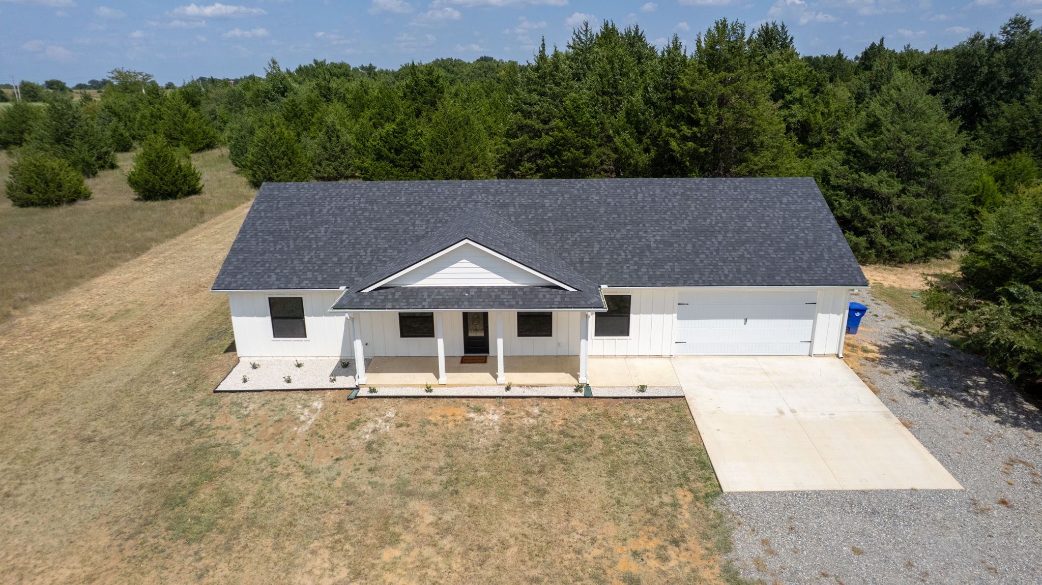 63 Deerfield Road Colbert, OK 74733 - Photo 29 of 33 Single-story property featuring a white exterior, dark gray shingle roof, covered front porch, an attached garage with a concrete driveway, and a gravel entry