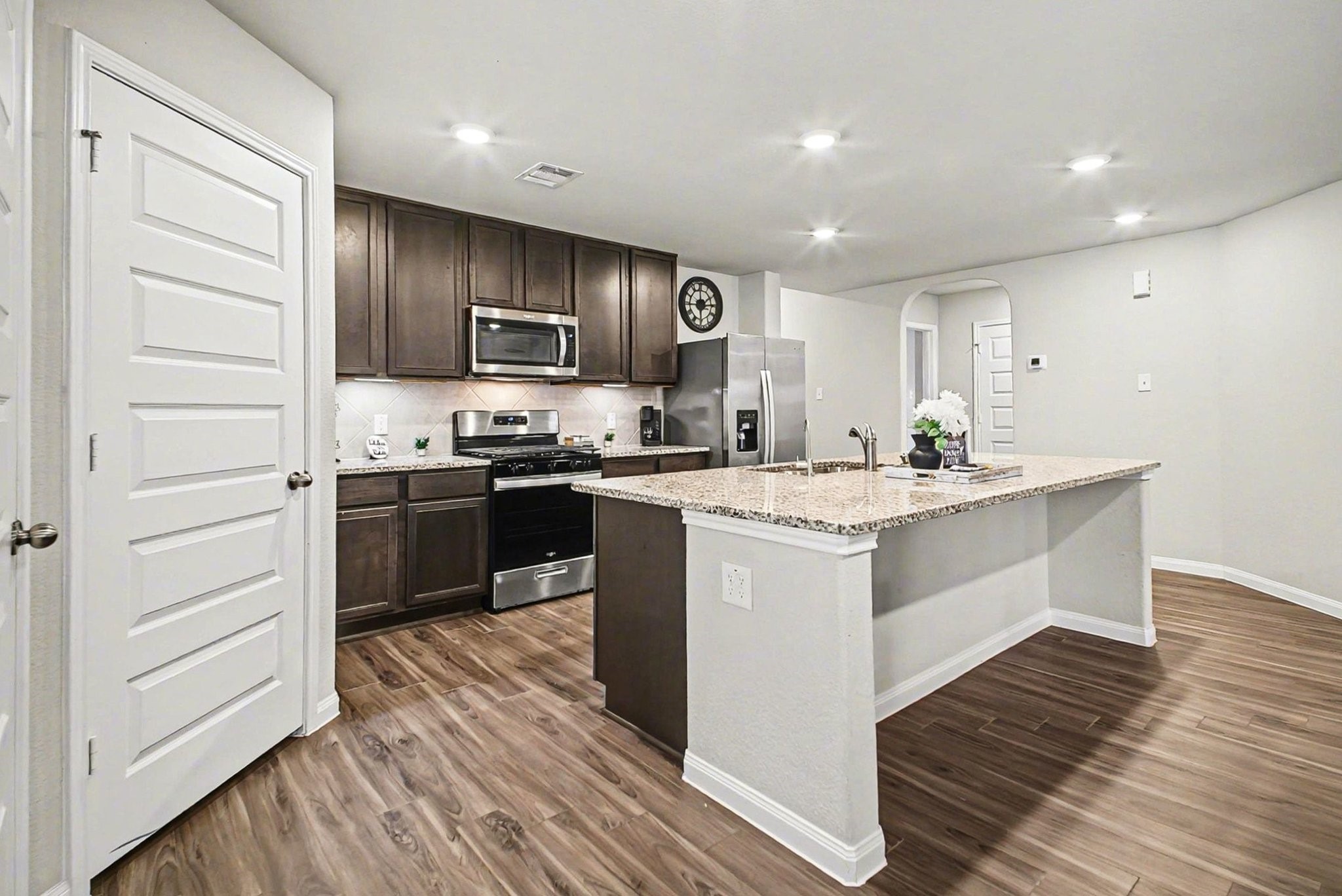4906 Luke Matthew Drive Rosharon, TX 77583 - Photo 8 of 34 This modern kitchen features dark wood cabinets, stainless steel appliances, and a spacious granite island with a sink. The open layout and recessed lighting enhance the bright, welcoming atmosphere, complemented by sleek wood flooring.
