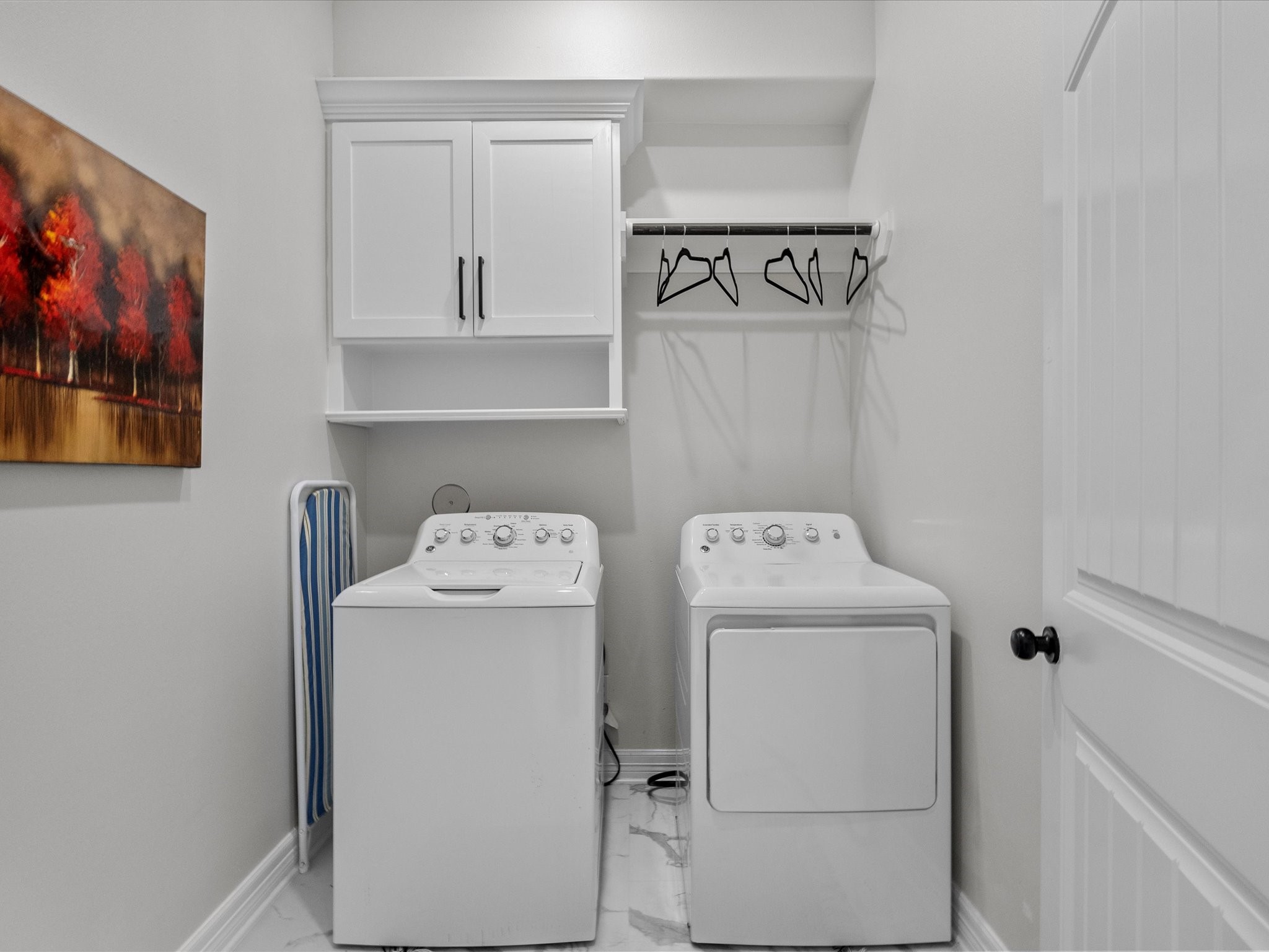 1018 Seashell Drive Surfside Beach, TX 77541 - Photo 33 of 49 This is a clean, organized laundry room featuring a washer and dryer, overhead cabinets, a hanging rod with hangers, and an ironing board. The room has a neutral color palette with a decorative wall painting.