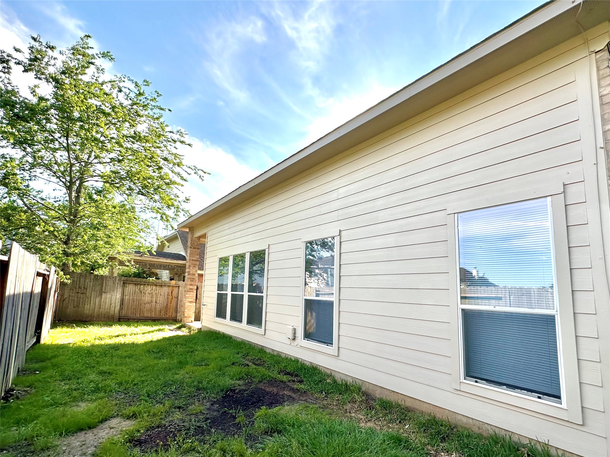 9611 Babbling Spring Court Tomball, TX 77375 - Photo 27 of 31 This photo shows a side view of a house with light-colored siding, featuring several large windows. There is a fenced-in yard with a grassy area and a tree, offering a cozy outdoor space.