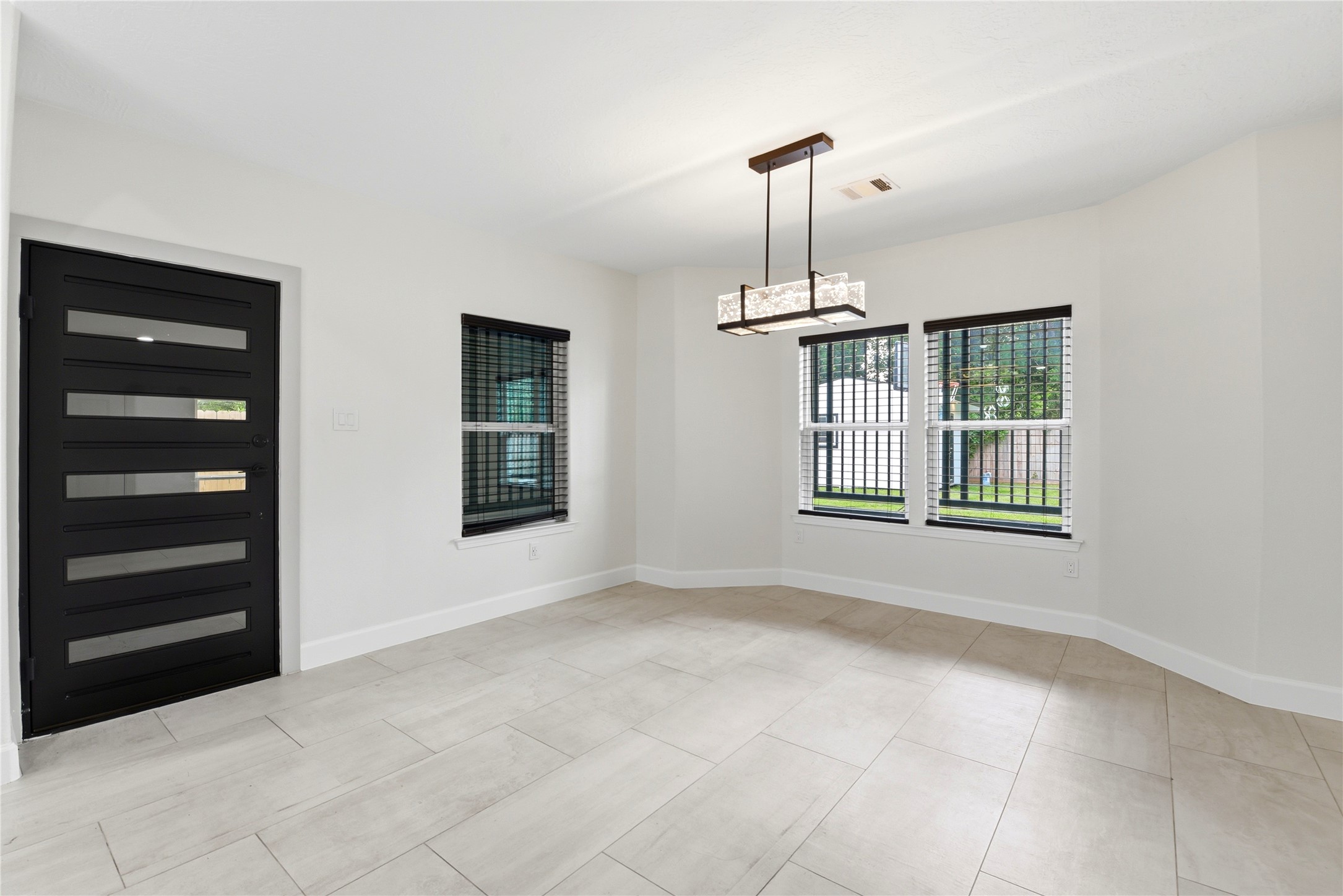 9403 Spaulding Street Houston, TX 77016 - Photo 16 of 50 This photo shows a bright, modern room with large windows, a sleek black door, and light-colored tile flooring. It features a contemporary rectangular light fixture, adding a stylish touch. Suitable for a dining or living area, its clean lines and neutral tones offer a versatile space.