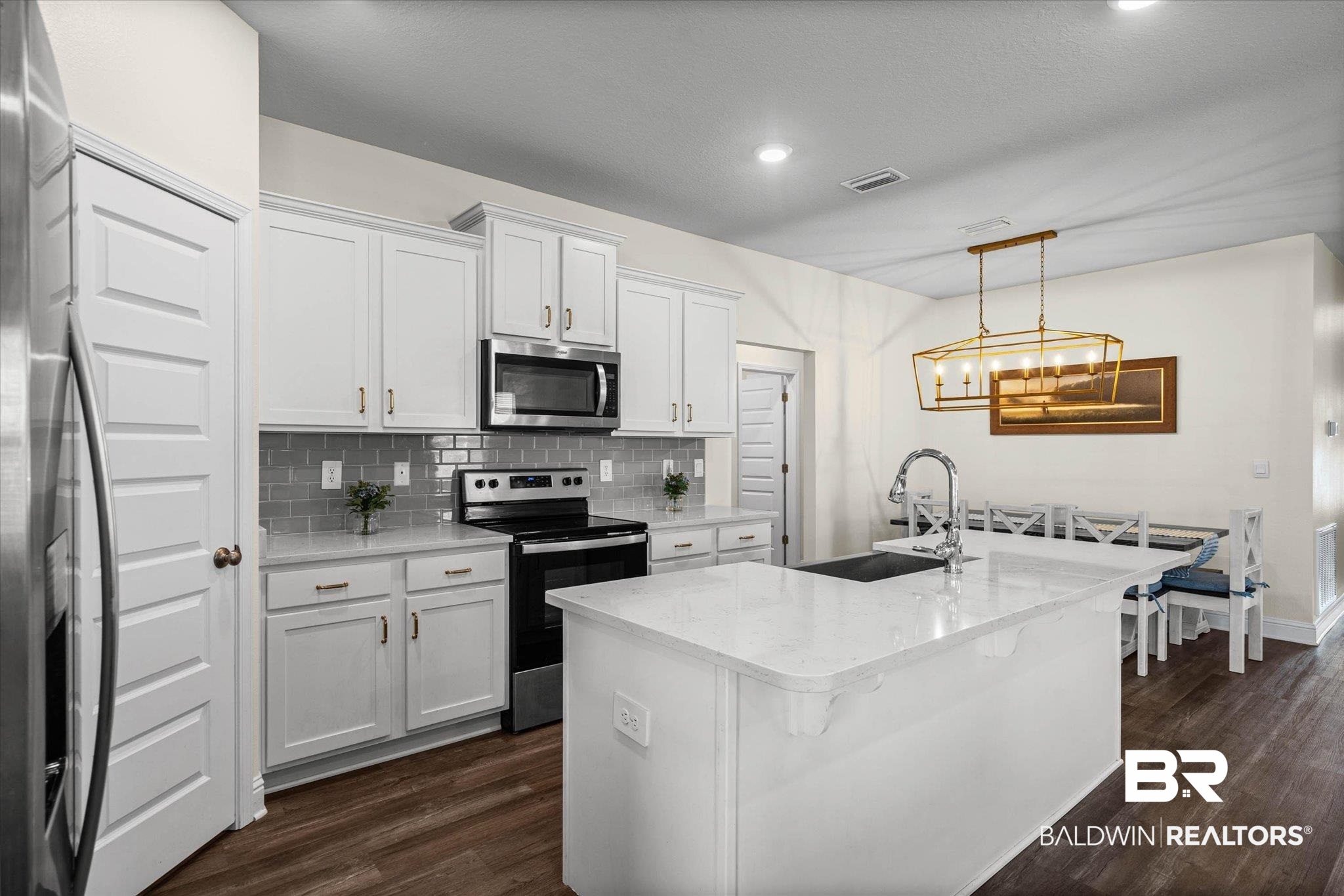 10541 Heirloom Road South Mobile, AL 36608 - Photo 8 of 23 Modern kitchen featuring a large central island with a sink, white cabinetry with gold-toned hardware, stainless steel appliances, grey subway tile backsplash, and wood-finish flooring