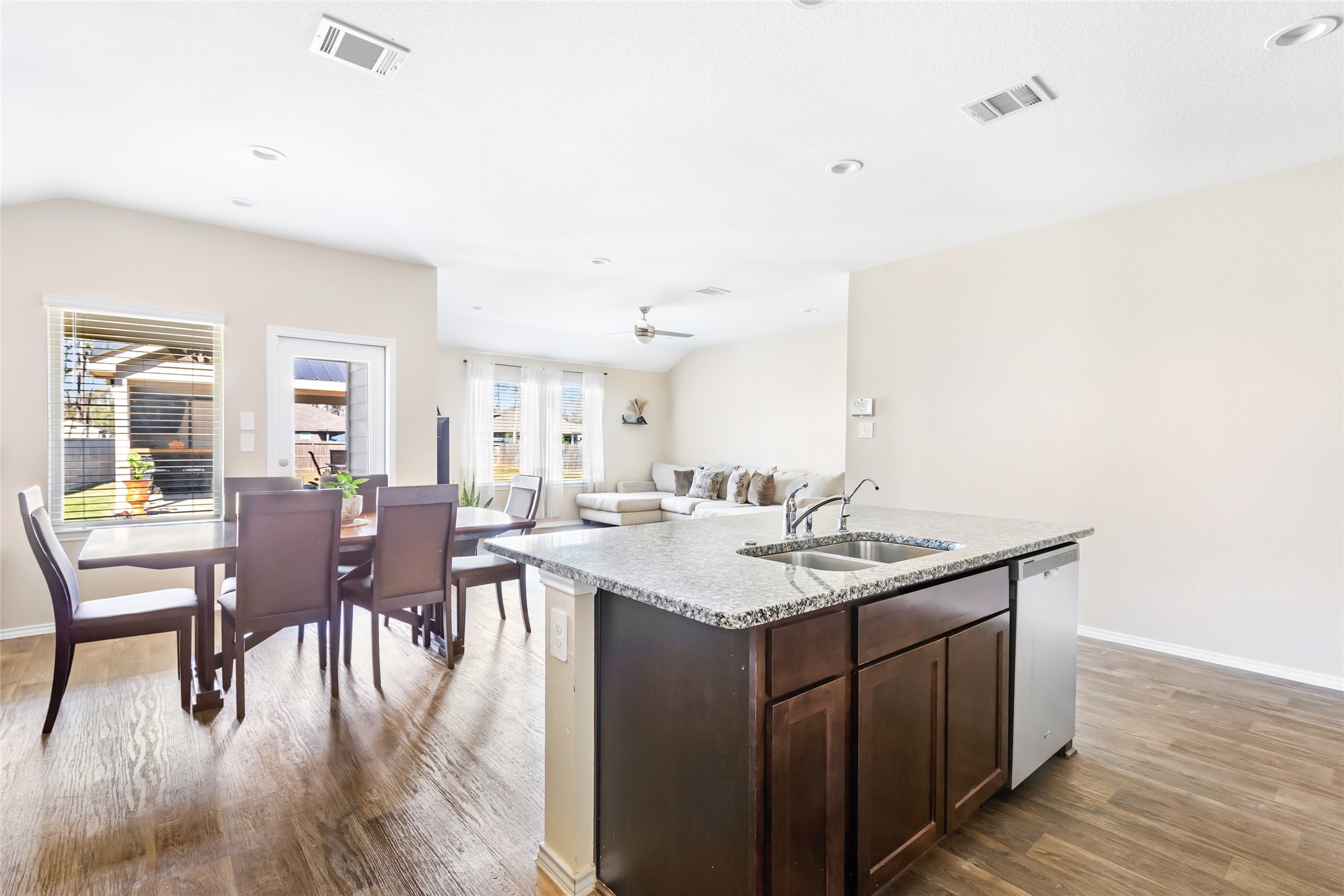 811 Republic Road New Caney, TX 77357 - Photo 26 of 37 This photo showcases a bright, open-plan living space with a modern kitchen island featuring granite countertops. The area includes a dining table with chairs and a cozy living room setup, highlighted by natural light from large windows and a glass door leading outside. The wood like flooring adds warmth to the contemporary design.