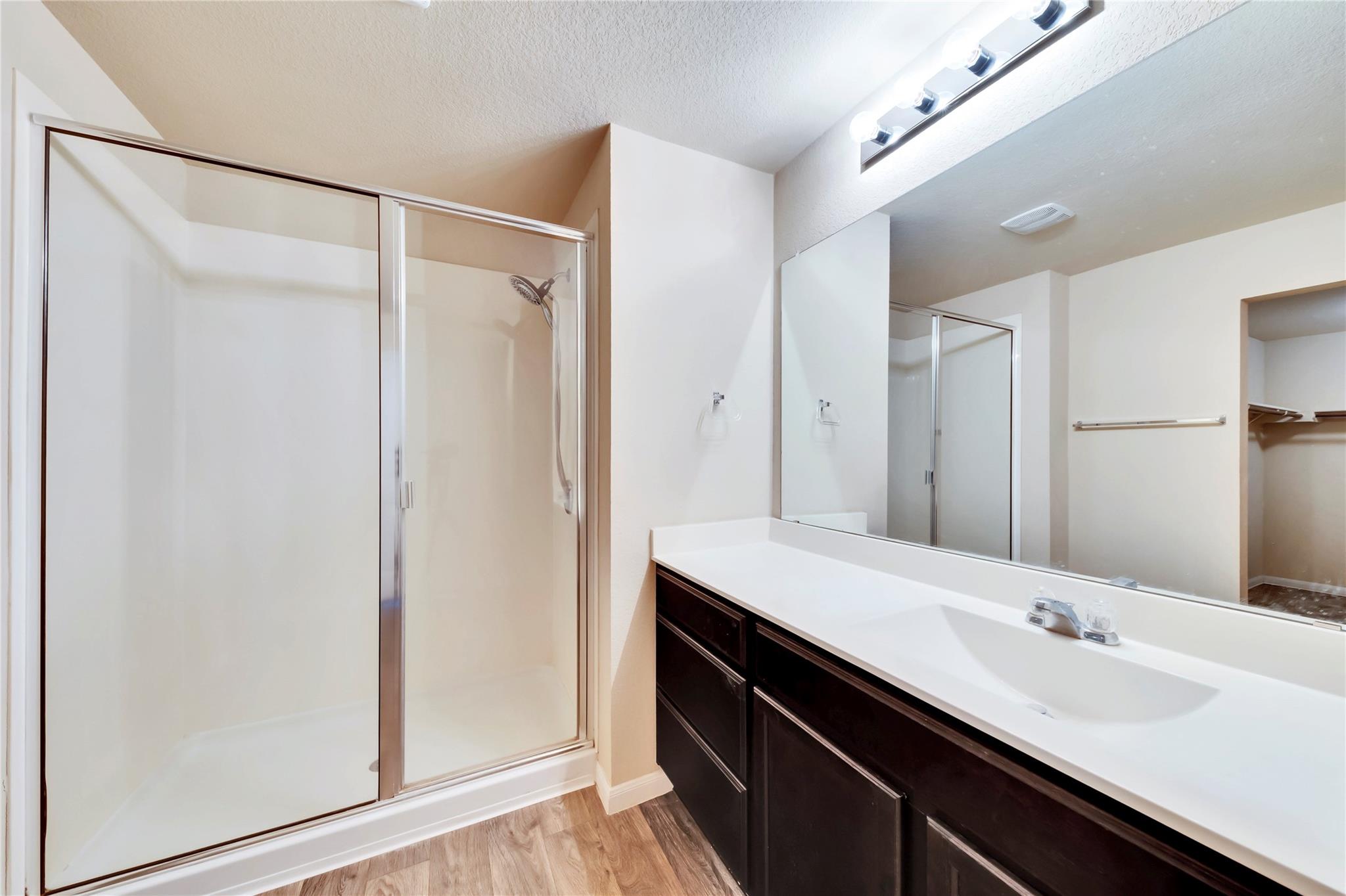 5913 Diamondleaf Bend Austin, TX 78724 - Photo 18 of 20 Bathroom featuring a walk-in shower with glass enclosure, a vanity with a white countertop and integrated sink, dark wood-finish cabinetry, a large wall-mounted mirror, and wood-finish flooring