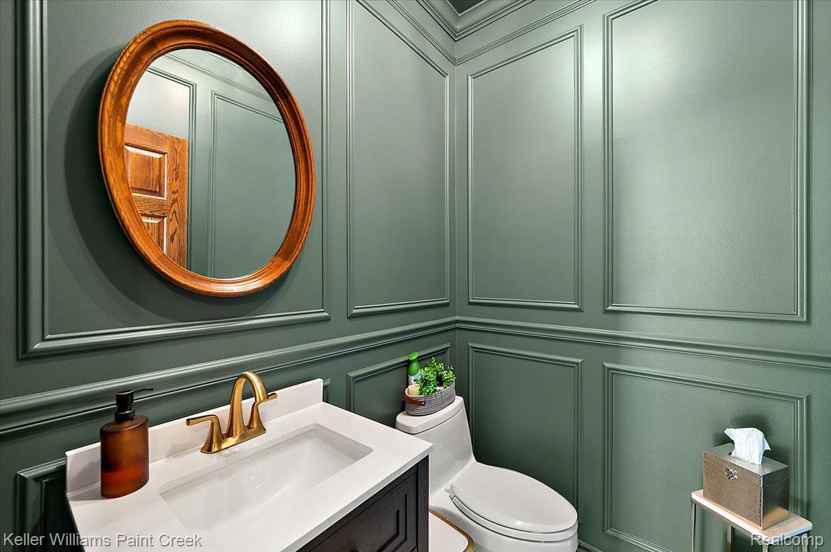 69069 Campground Road Romeo, MI 48065 - Photo 38 of 78 Powder room featuring detailed paneled walls, a white countertop vanity with a gold-finish faucet, and a round wood-framed mirror