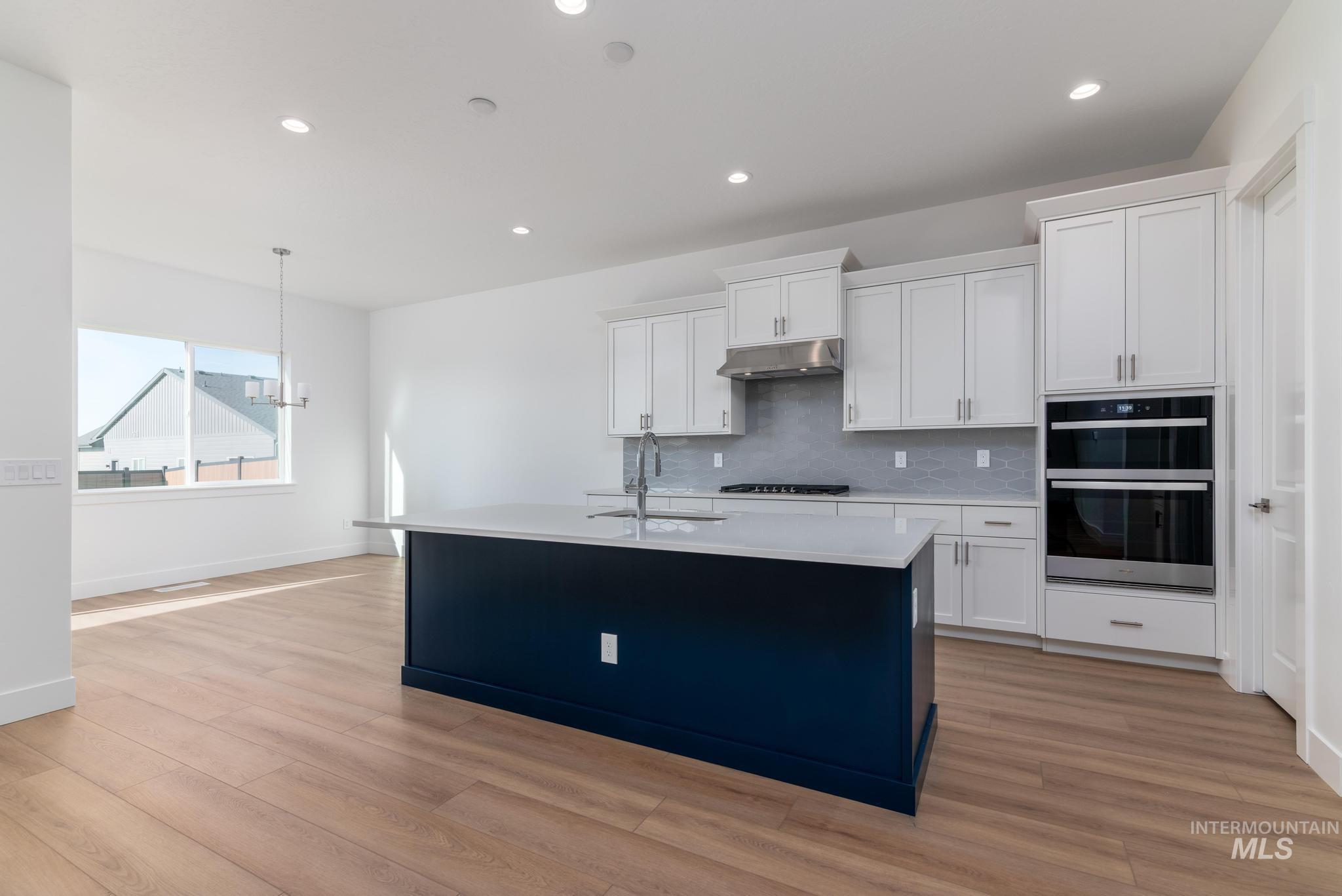 5403 Juliet Rose Avenue Caldwell, ID 83607 - Photo 4 of 12 Modern kitchen featuring a contrasting navy island with countertop seating, light wood-finish flooring, white shaker cabinetry, stainless steel appliances including a range hood and double wall ovens, and recessed ceiling lighting