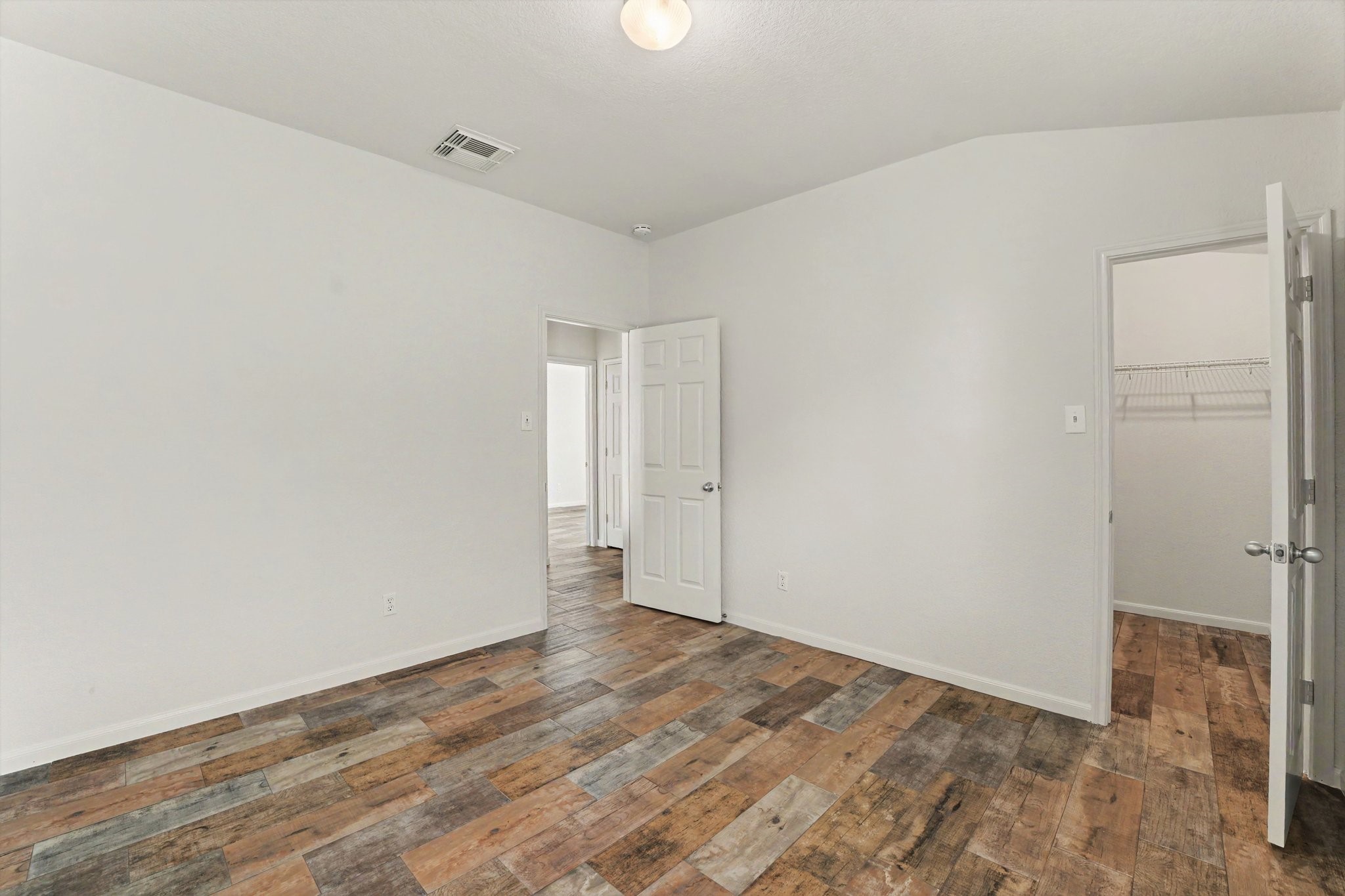 21427 Blooming Rock Lane Spring, TX 77379 - Photo 27 of 32 This photo shows a bright, empty room with light-colored walls and rustic wood-look flooring. There are two doors: one leading to a hallway and another to a walk-in closet. The ceiling features a simple light fixture.