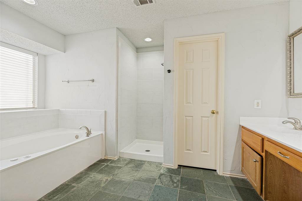 1215 Spring Creek Drive Allen, TX 75002 - Photo 17 of 40 Bathroom featuring a spacious soaking tub, separate shower stall with white tile surround, a single vanity with wood-finish cabinetry, a white countertop, and dark slate flooring
