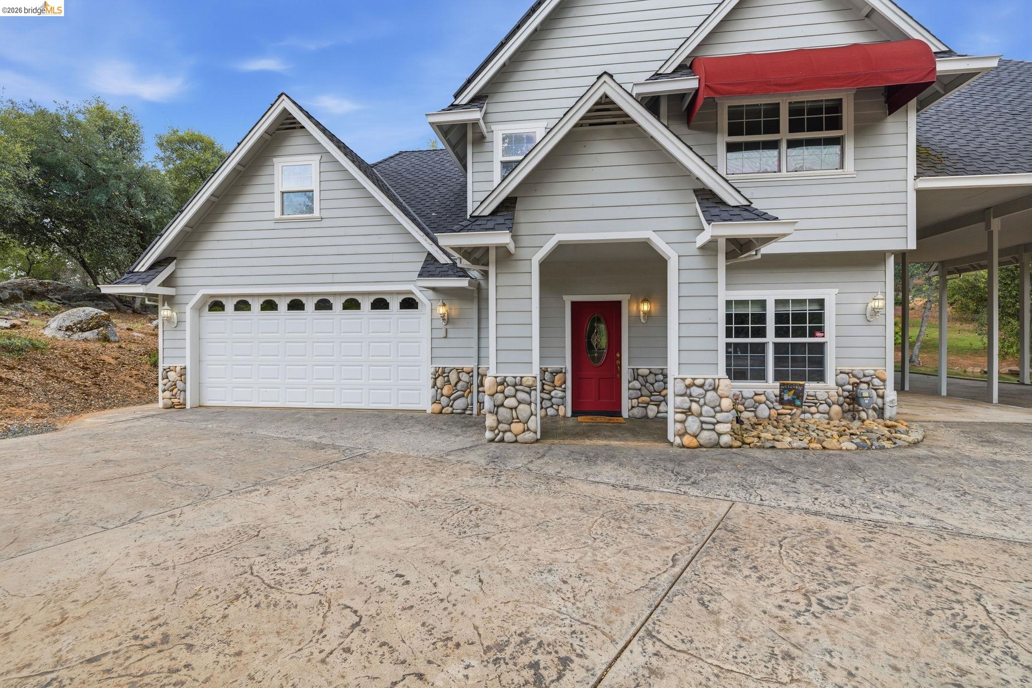 18450 Lambert Lake Road Sonora, CA 95370 - Photo 8 of 87 Grey siding exterior with stone accents, a red entry door, an attached two-car garage, and a covered carport