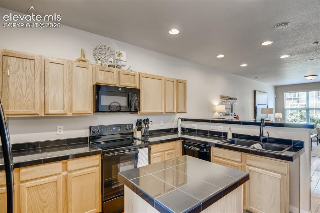 846 Victoria Ridge Point Colorado Springs, CO 80903 - Photo 9 of 27 Kitchen featuring light wood cabinetry, black tile countertops, black appliances, a central island with black tile top, and recessed ceiling lighting