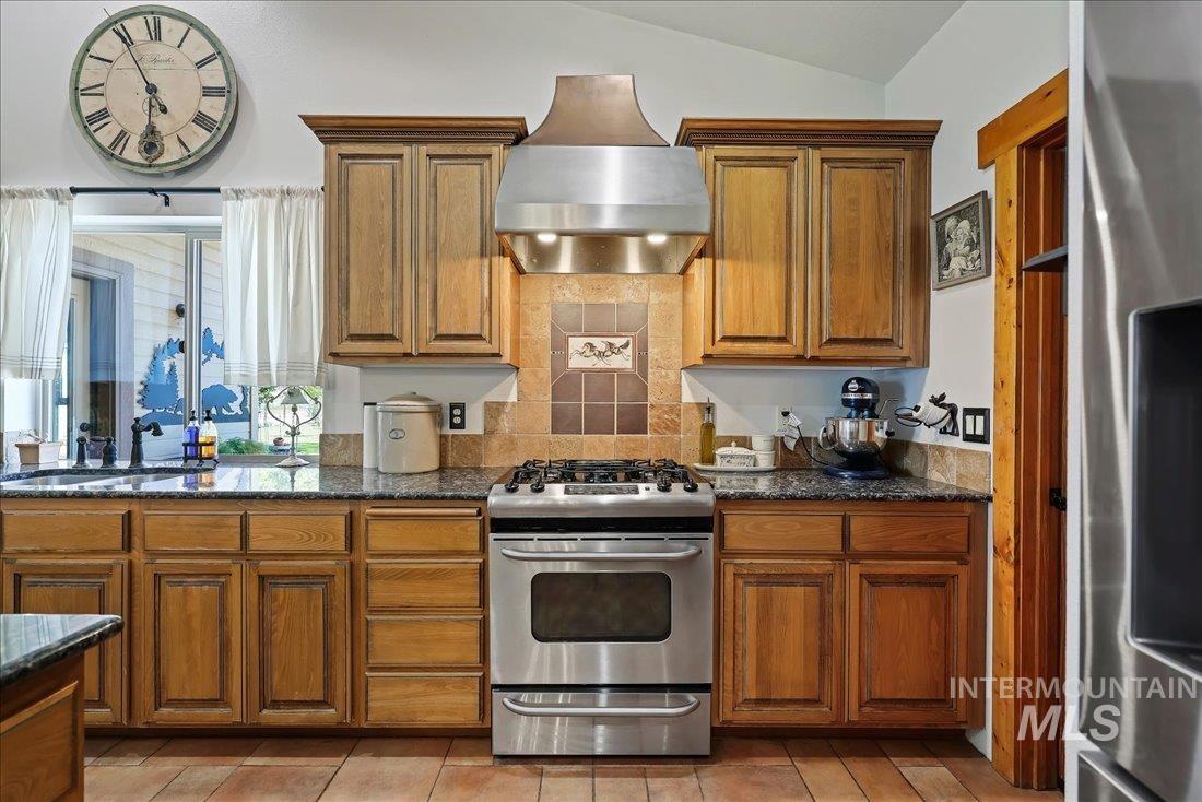 7639 Elko Lane Middleton, ID 83644 - Photo 13 of 47 Kitchen with stainless steel appliances, wall chimney range hood, brown cabinetry, dark stone countertops, and lofted ceiling