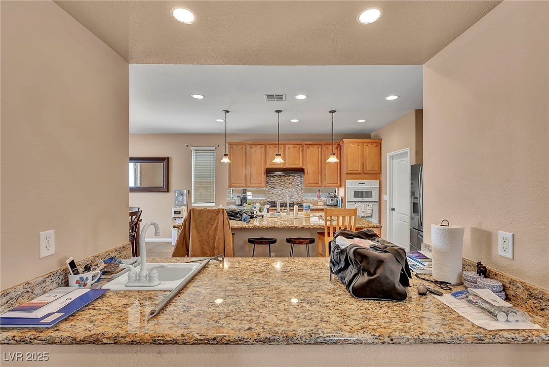 8208 Seven Falls Street North Las Vegas, NV 89085 - Photo 12 of 69 Kitchen featuring backsplash, a peninsula, light stone countertops, pendant lighting, and stainless steel fridge with ice dispenser