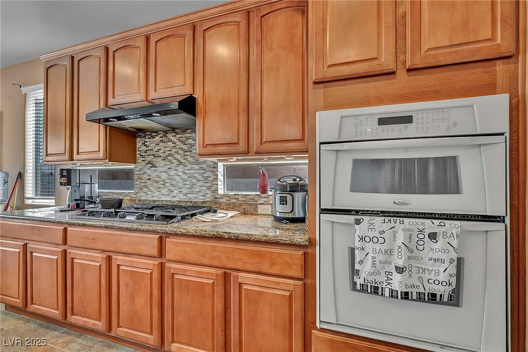 8208 Seven Falls Street North Las Vegas, NV 89085 - Photo 8 of 69 Kitchen with double oven, decorative backsplash, under cabinet range hood, light stone counters, and stainless steel gas stovetop