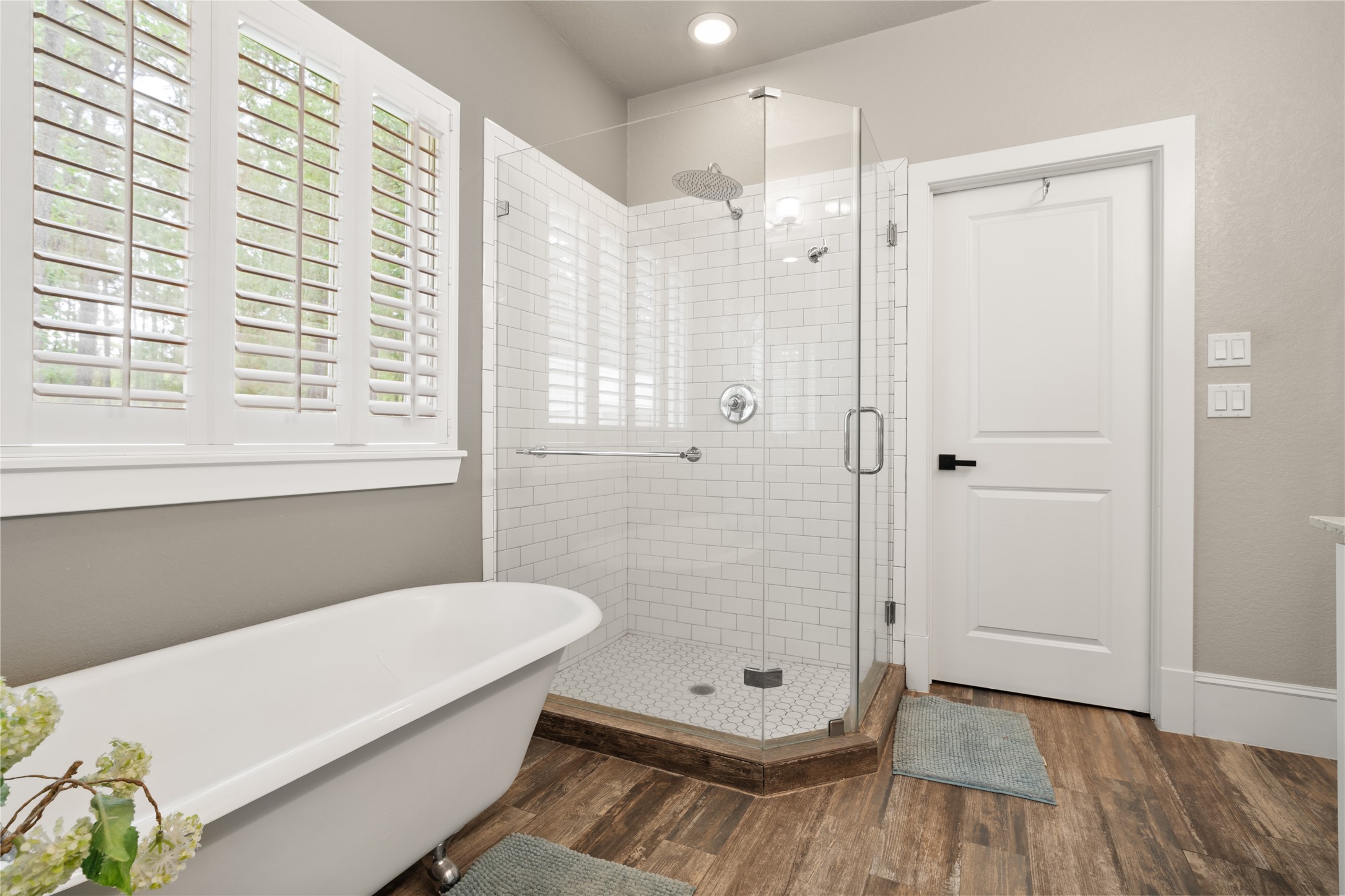 36 Wind River Drive Conroe, TX 77384 - Photo 30 of 45 This photo showcases a modern bathroom featuring a standalone clawfoot tub, a spacious glass-enclosed shower with subway tile, and elegant wood-look flooring. The room is bright, with large windows covered by plantation shutters, offering a clean and inviting atmosphere.