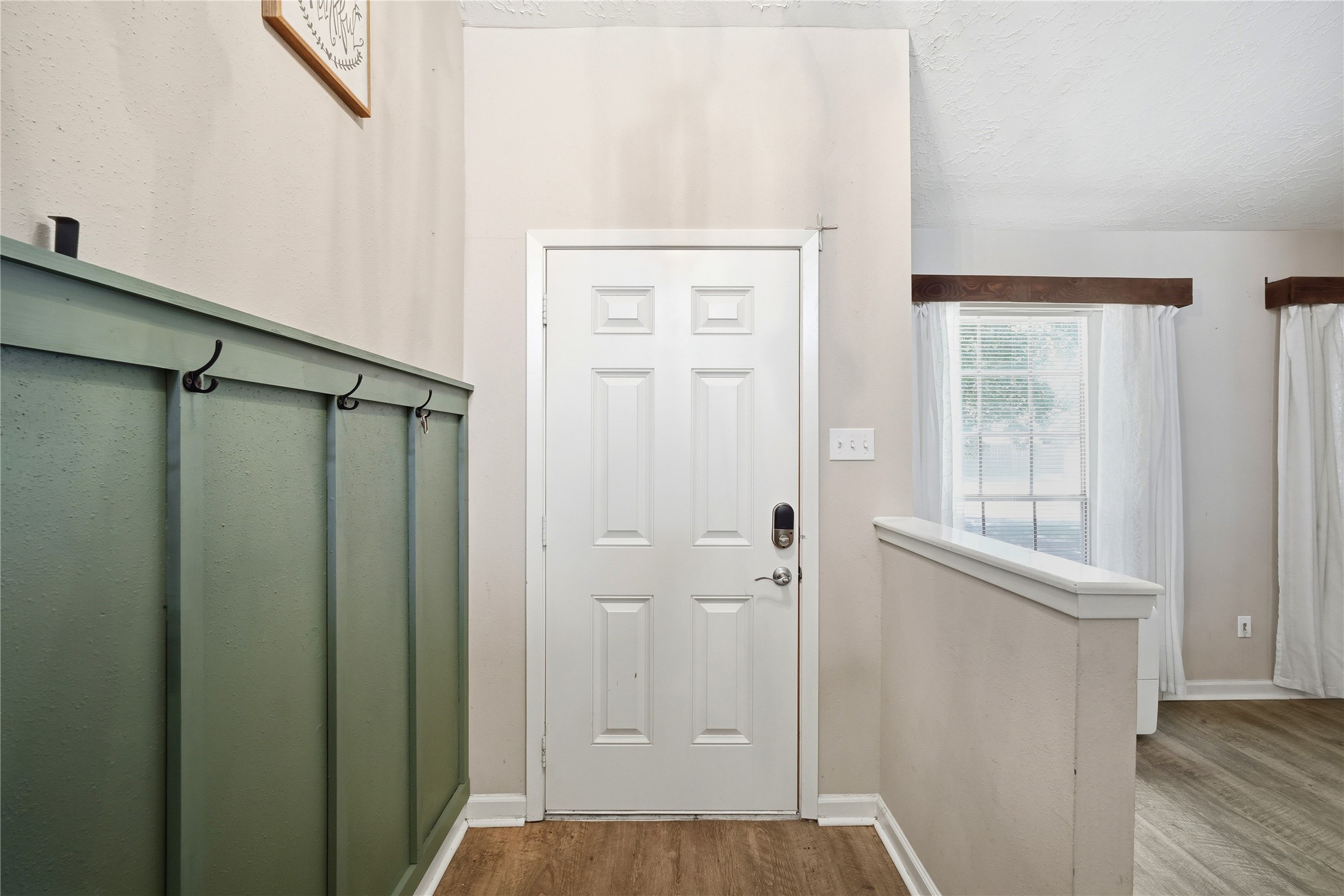 4718 Temple Bell Drive Spring, TX 77388 - Photo 6 of 50 This photo shows a welcoming entryway with a white front door, modern keypad lock, and hooks for hanging items. A green accent wall adds character, and natural light streams in from a window with white curtains, enhancing the warm wood laminate flooring.