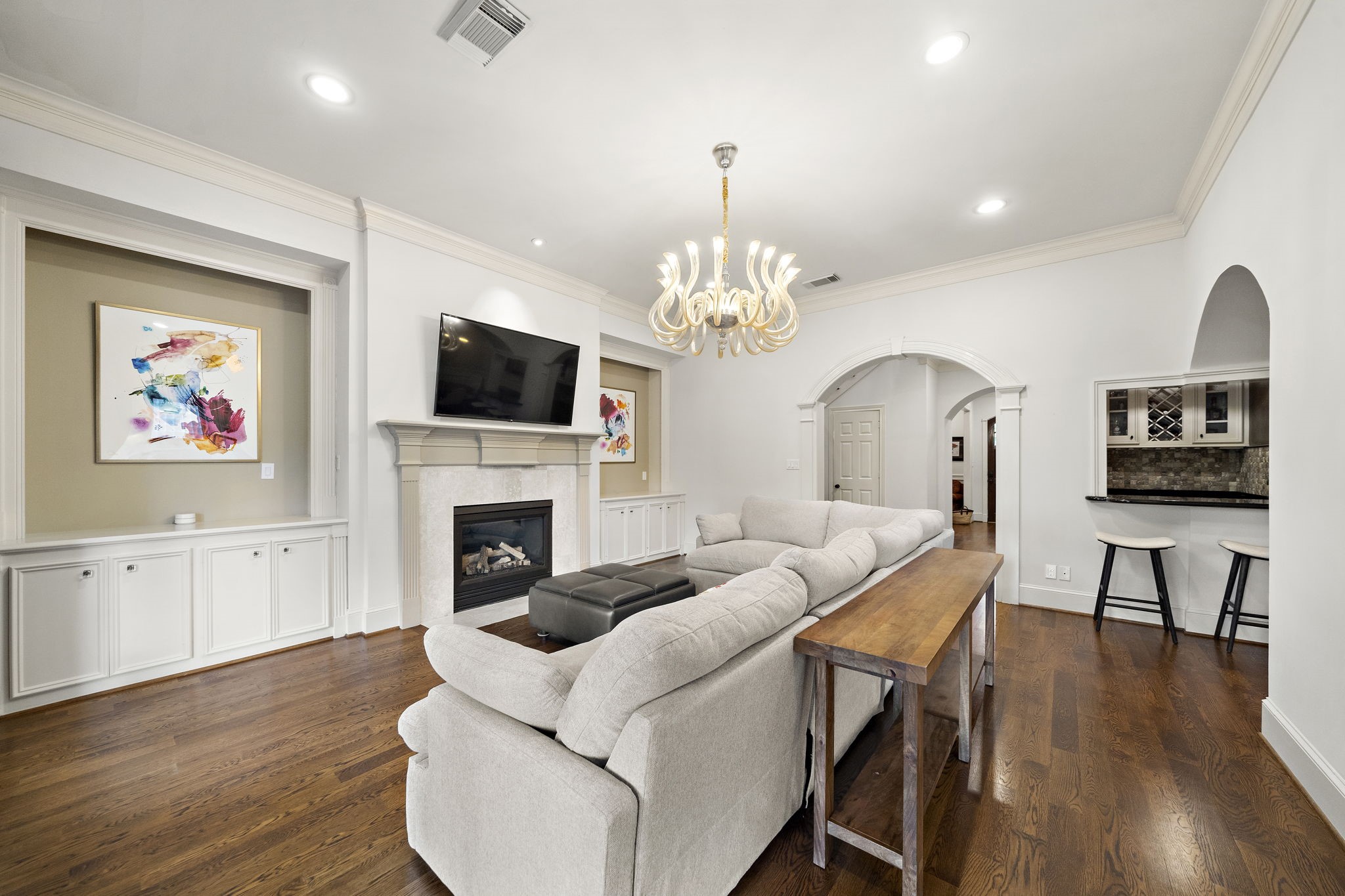 1229 Ripple Creek Drive Houston, TX 77057 - Photo 12 of 49 This living room features elegant hardwood floors, a cozy fireplace, and built-in cabinetry. It boasts a modern chandelier, a wall-mounted TV, and an open layout with archways leading to additional spaces. A small bar area with seating adds a touch of luxury.