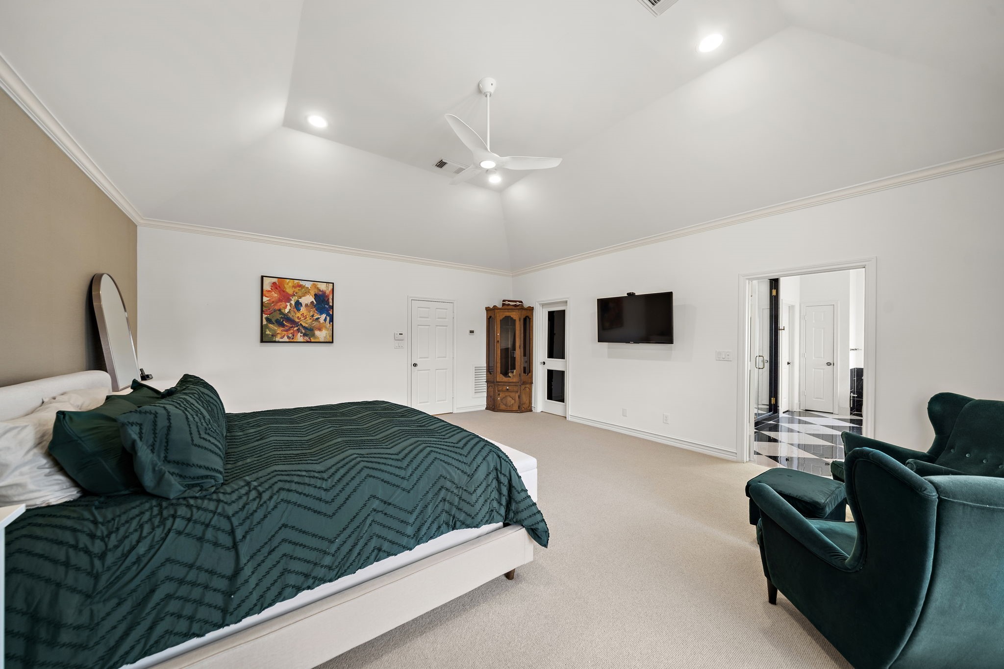 1229 Ripple Creek Drive Houston, TX 77057 - Photo 24 of 49 Spacious bedroom with high ceilings and recessed lighting. Features a large bed with green bedding, an armchair, wall-mounted TV, and an adjoining bathroom with a checkered floor. Neutral tones and modern decor create a cozy atmosphere.